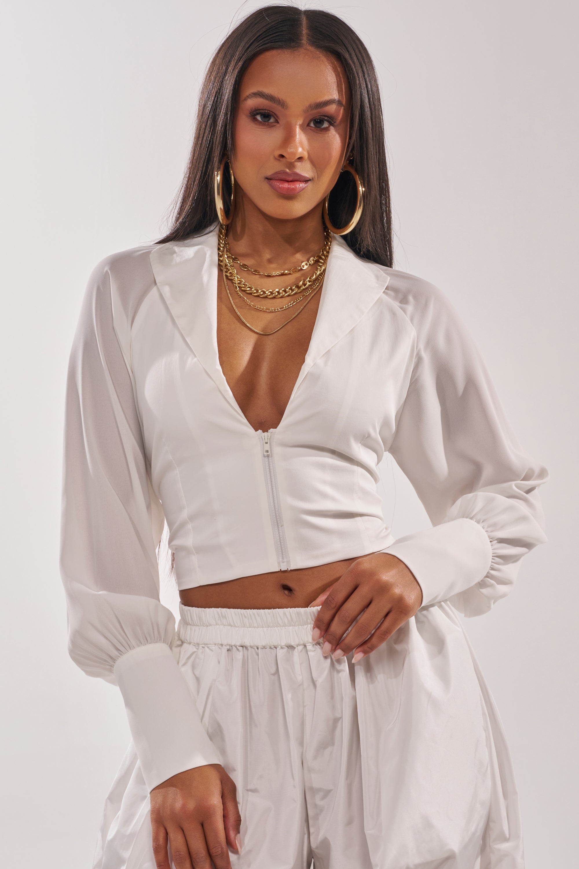 A woman with long hair wears the EFFORTLESS CORSET BLOUSE TOP in white, styled with matching high-waisted pants, gold hoop earrings, and layered gold necklaces. She stands against a plain light background. 1