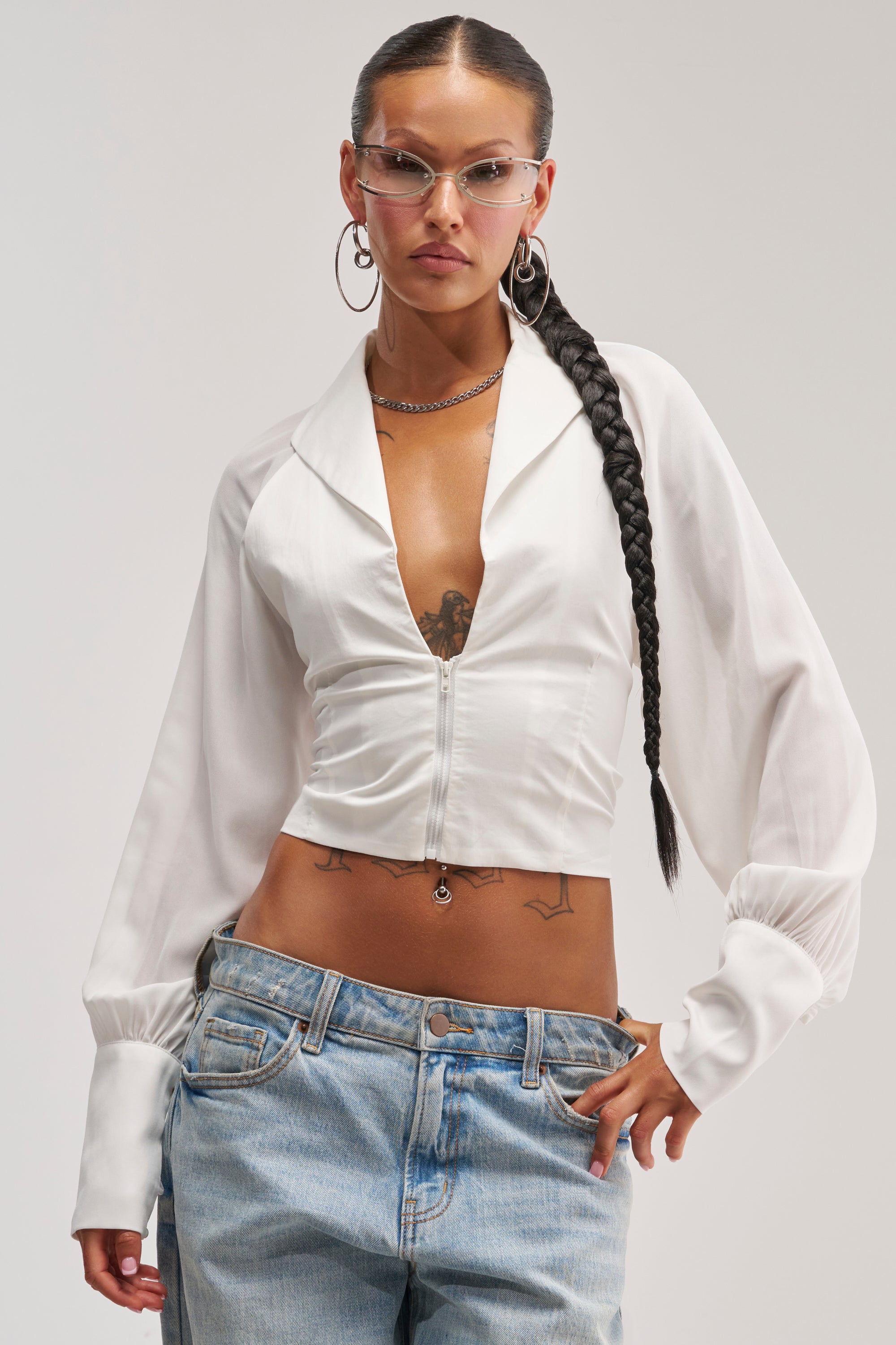 A woman with a long braid wears glasses, hoop earrings, and the EFFORTLESS CORSET BLOUSE TOP featuring a front zipper, styled with light blue jeans. Displaying tattoos on her chest and arms, she poses confidently against a plain background.