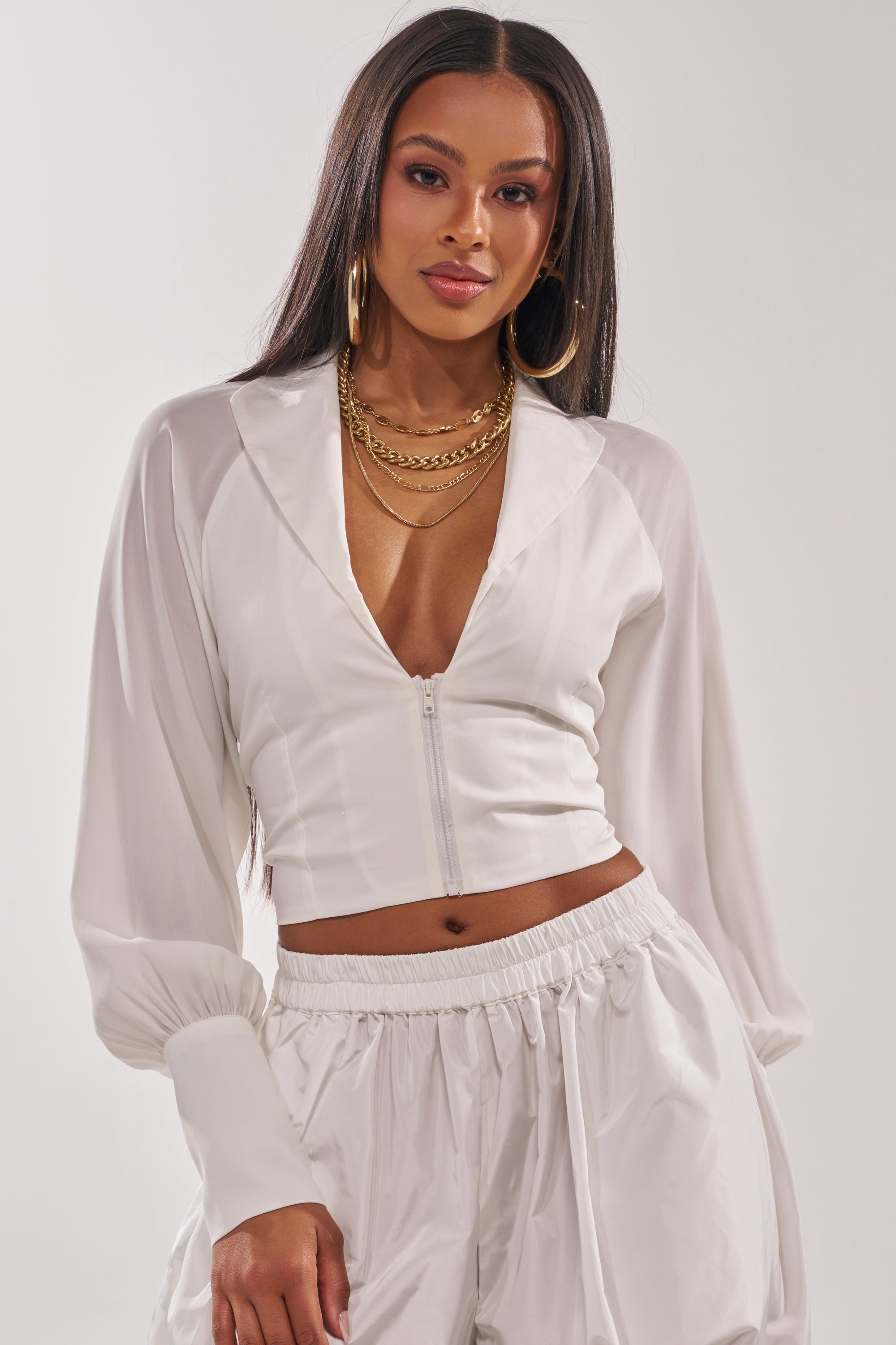 A woman showcases the EFFORTLESS CORSET BLOUSE TOP, a white long sleeve zip-up crop with a deep V-neck, styled with matching high-waisted pants and finished with layered gold necklaces and hoop earrings for a chic, coordinated look. 2