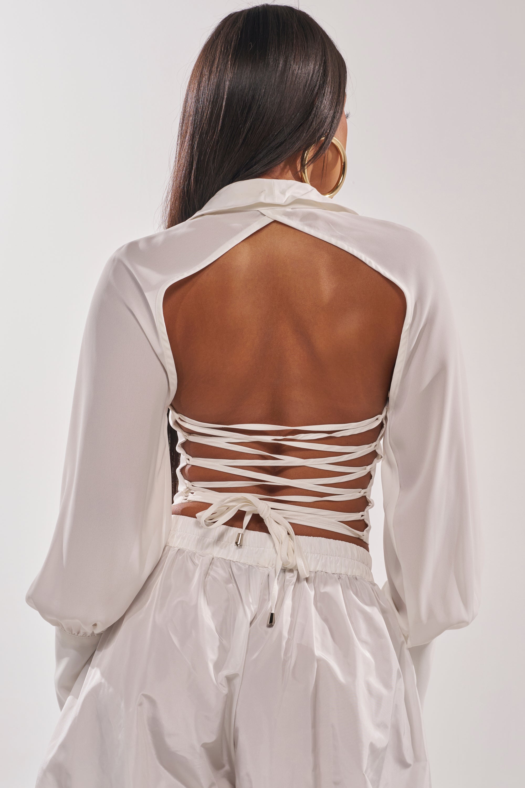 A woman with long dark hair models the EFFORTLESS CORSET BLOUSE TOP, featuring a white backless, crisscross lace-up design and loose sleeves, paired with high-waisted white pants as she stands with her back to the camera. 4