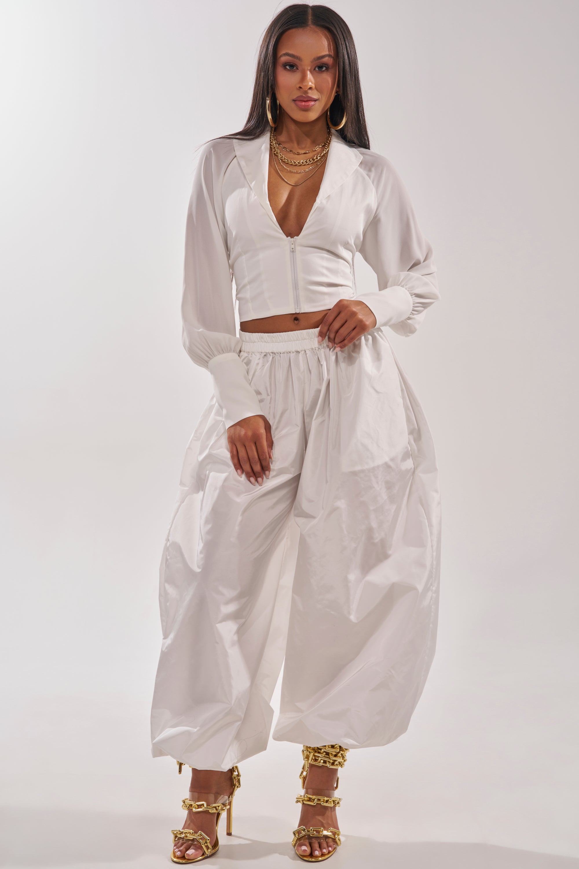 A woman exudes confidence in the EFFORTLESS CORSET BLOUSE TOP, a chic white zip-up blouse paired with loose pants, gold chain necklaces, and gold high-heeled sandals against a plain light background. 5