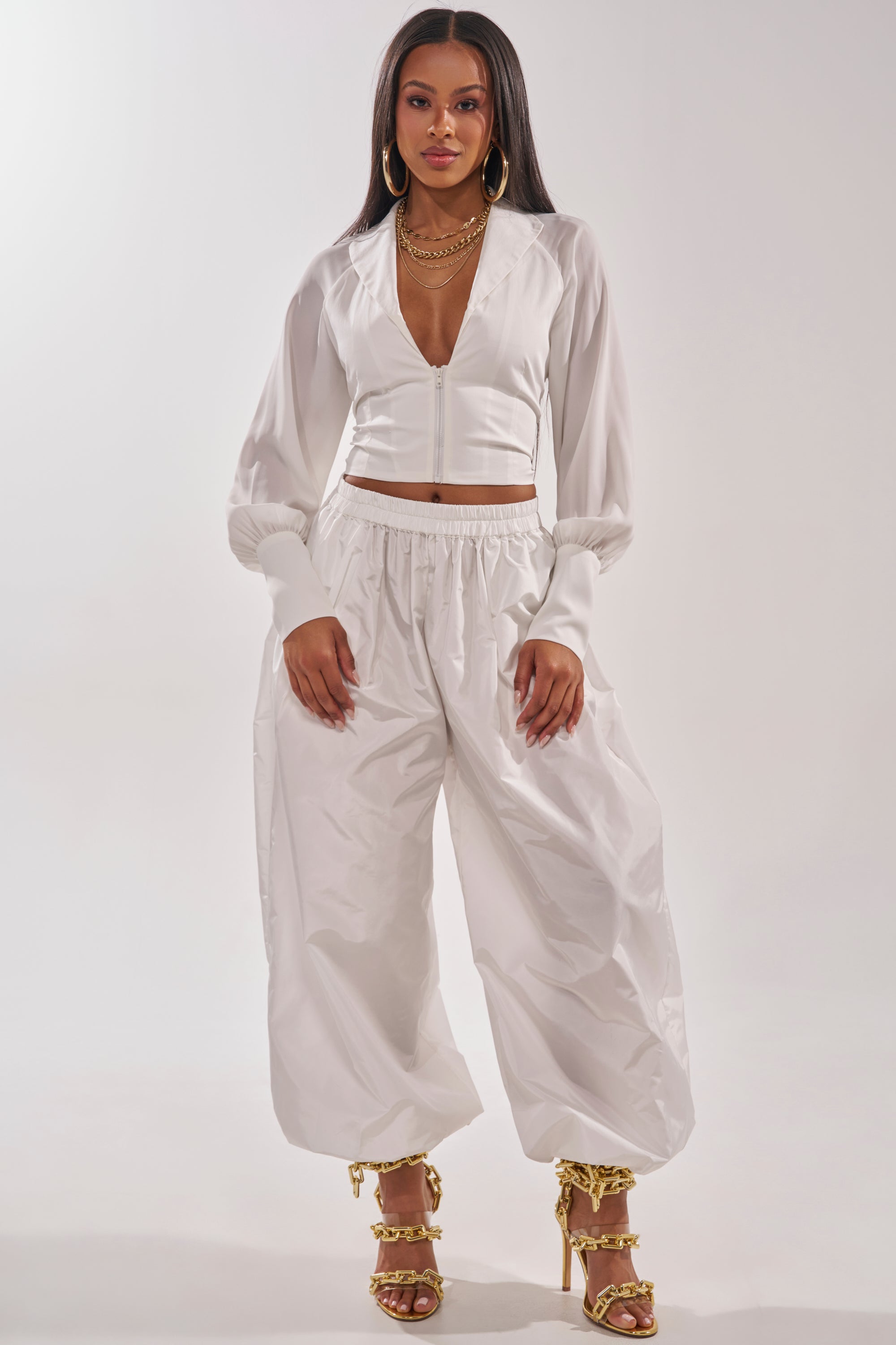A woman stands confidently in the EFFORTLESS CORSET BLOUSE TOP, styled as a crop top with loose white pants and gold high-heeled sandals. She completes the look with gold hoop earrings and layered necklaces against a plain background. 6