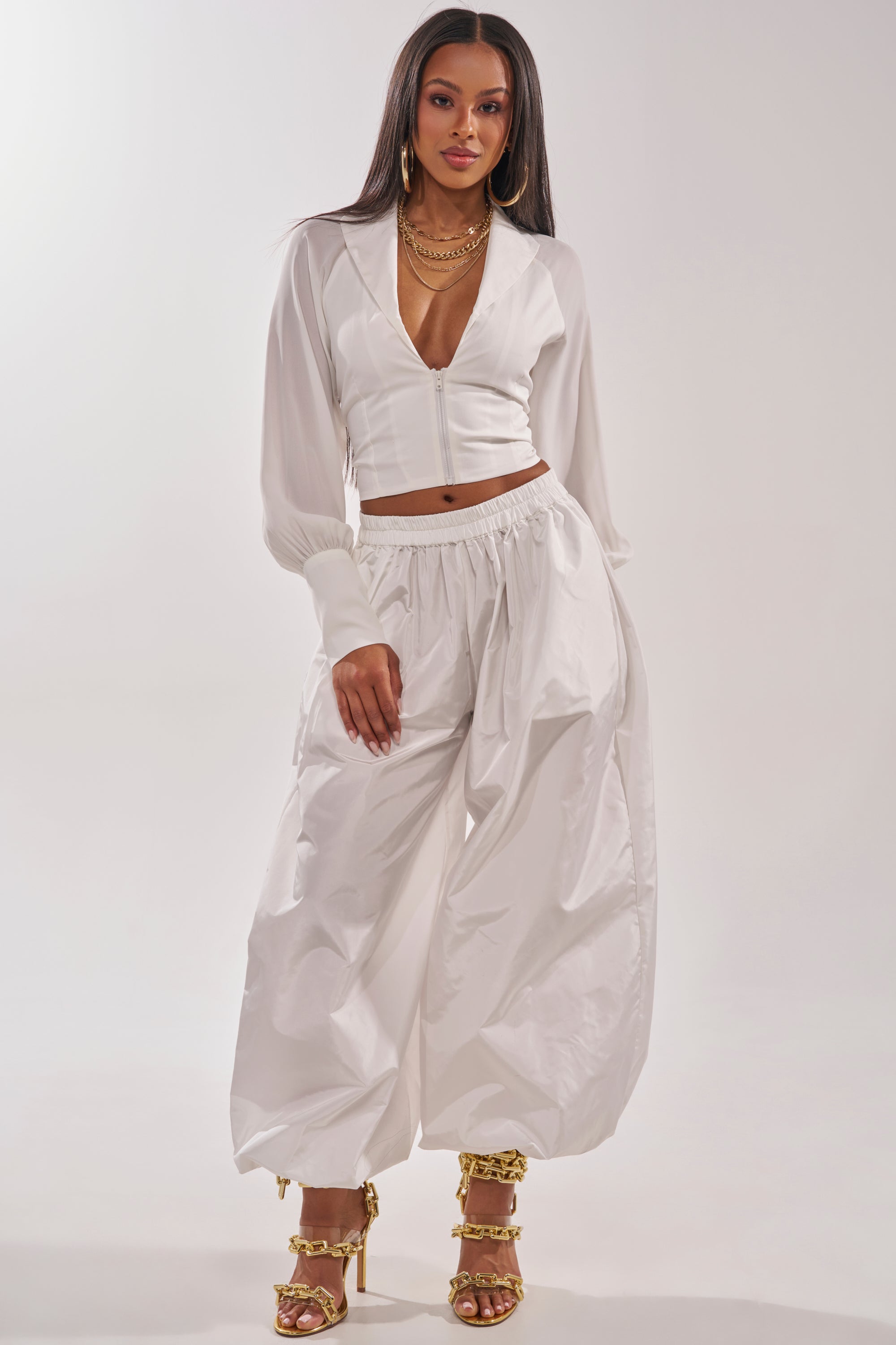 A woman stands confidently in an EFFORTLESS CORSET BLOUSE TOP, paired with loose high-waisted white pants, gold high-heeled sandals, and layered gold jewelry against a light background. 7