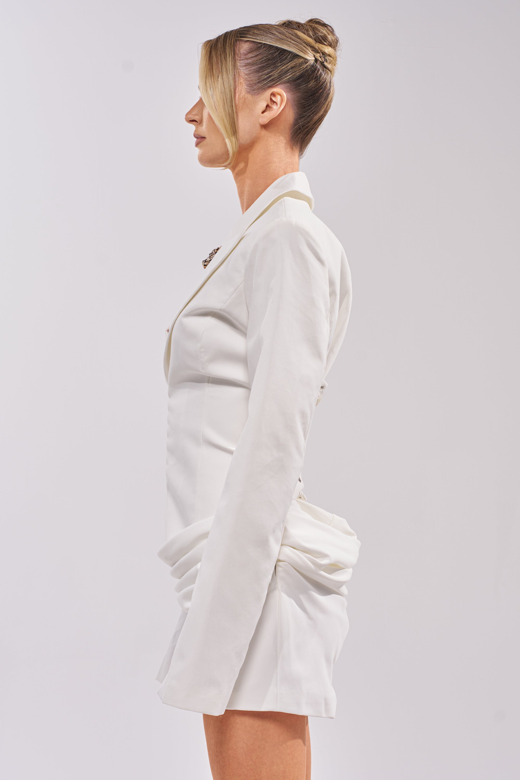A woman in profile stands against a plain background, wearing the BUSINESS IN THE FRONT CORSET BLAZER MINI DRESS—white with a corseted waist and asymmetric draped skirt. Her blonde hair is styled in a sleek updo as she faces left. 2