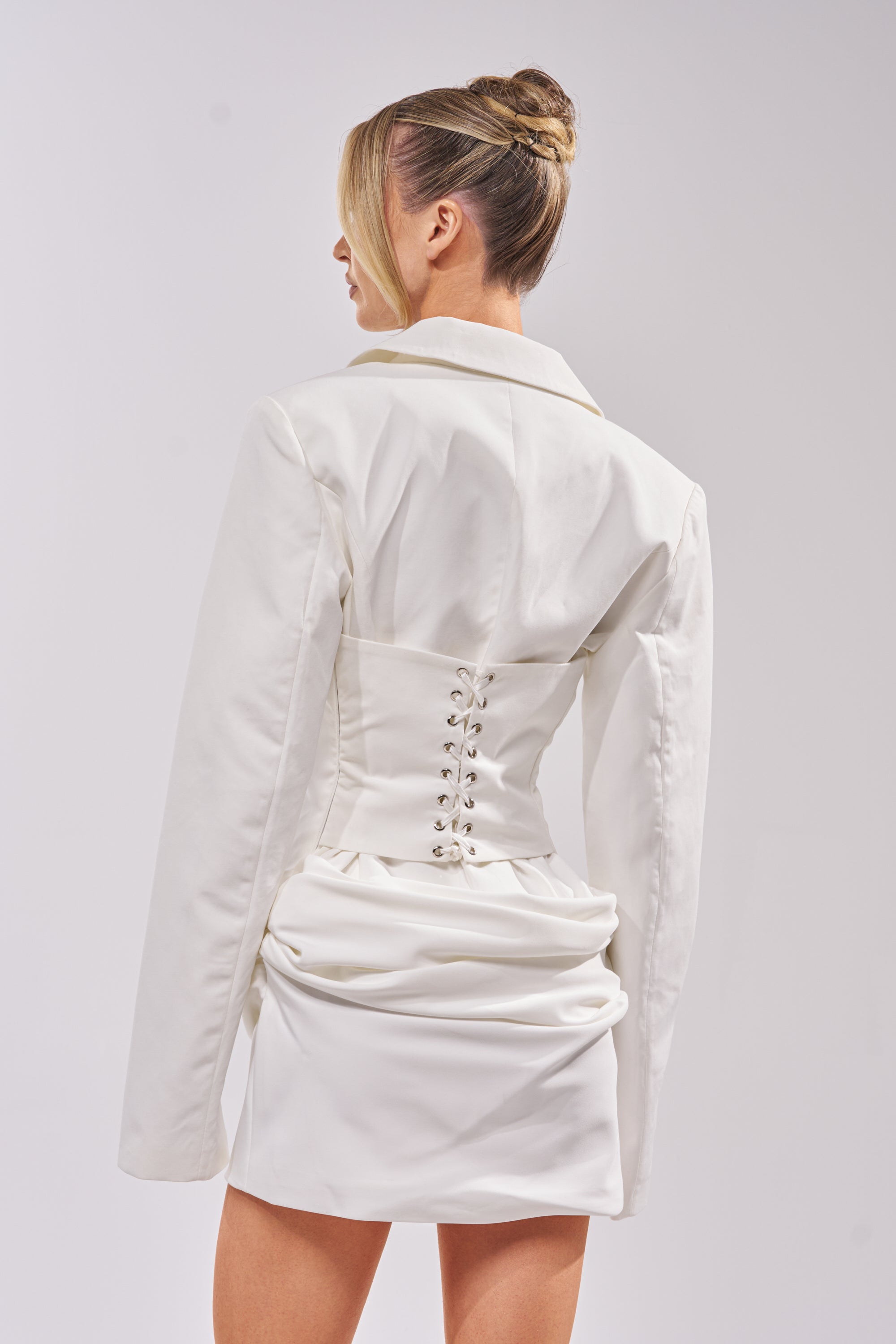 A woman with her hair in a neat bun faces away, modeling the BUSINESS IN THE FRONT CORSET BLAZER MINI DRESS, featuring a white corseted blazer style, lace-up back detail, and short skirt against a plain background. 3