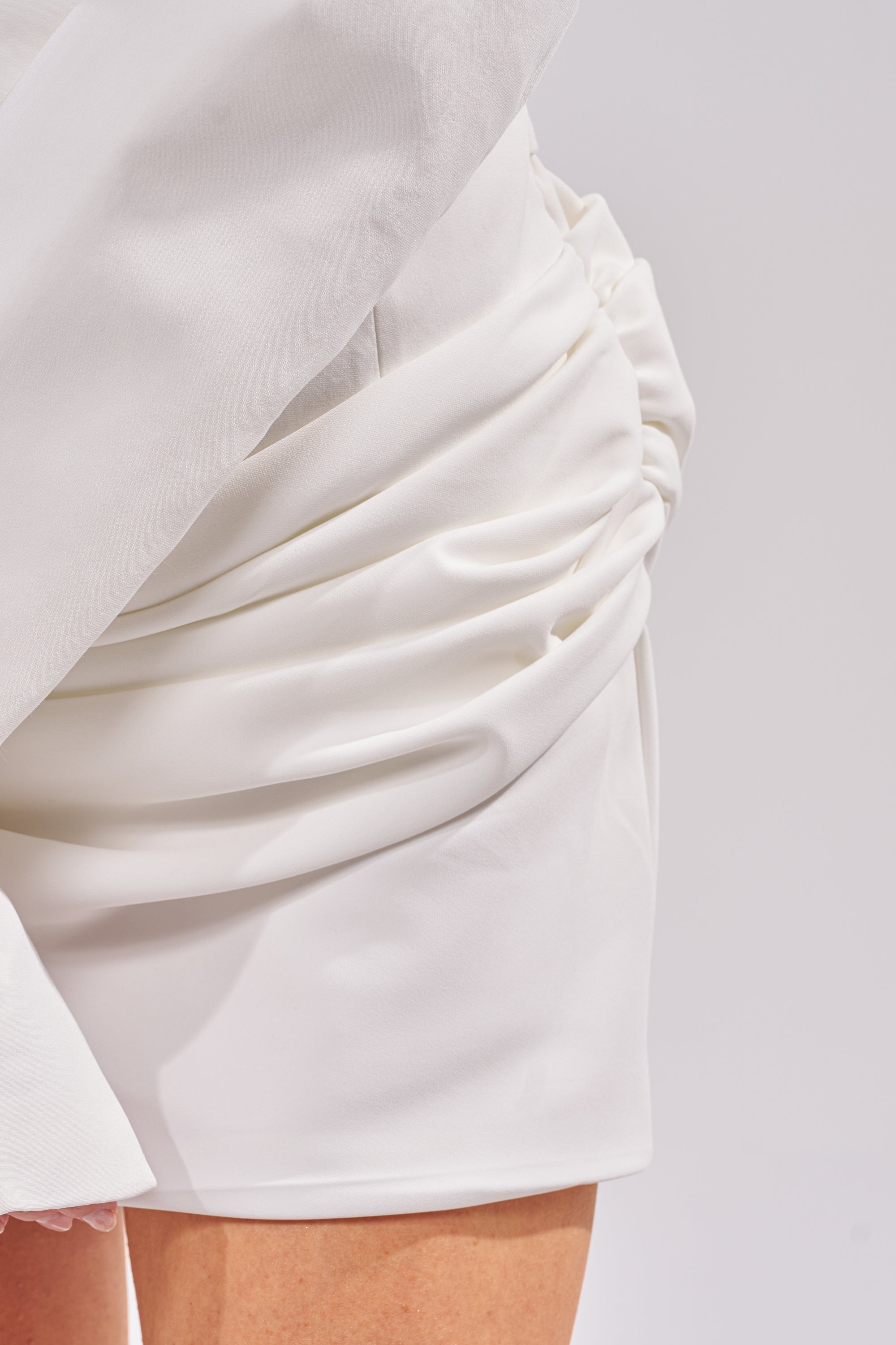 Close-up of a person wearing the BUSINESS IN THE FRONT CORSET BLAZER MINI DRESS, featuring white ruched and draped fabric that reveals part of their bare leg against a plain, light-colored background. 7