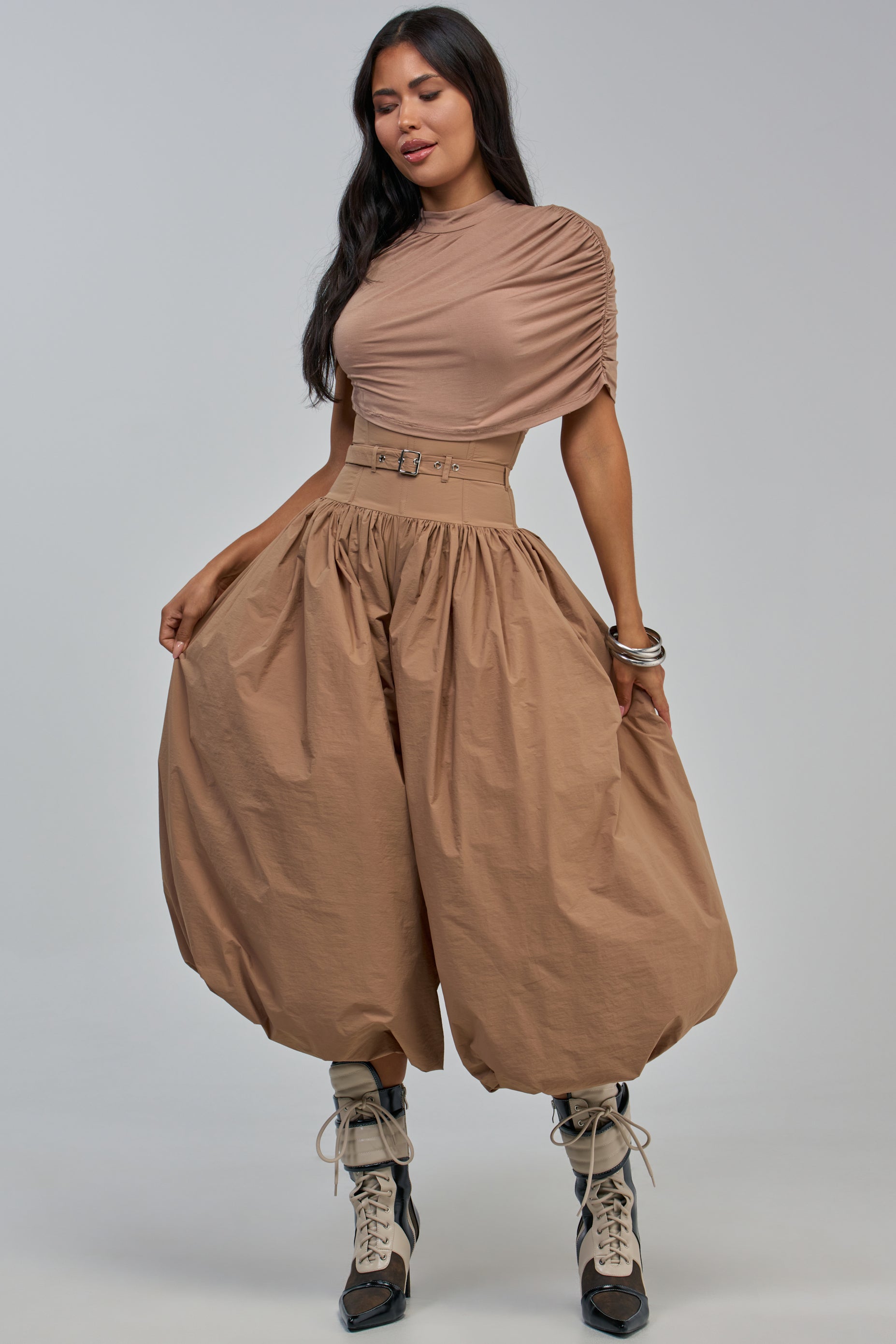 Wearing the “SPIN ME AROUND POPLIN JUMPSUIT IN TAUPE,” a sophisticated ensemble featuring ruched sleeves, wide high-waisted balloon legs, and a belted waist in a chic taupe hue. Styled with lace-up boots and silver bangles against a plain background, this outfit is perfect for special occasions, blending modern flair with timeless elegance.