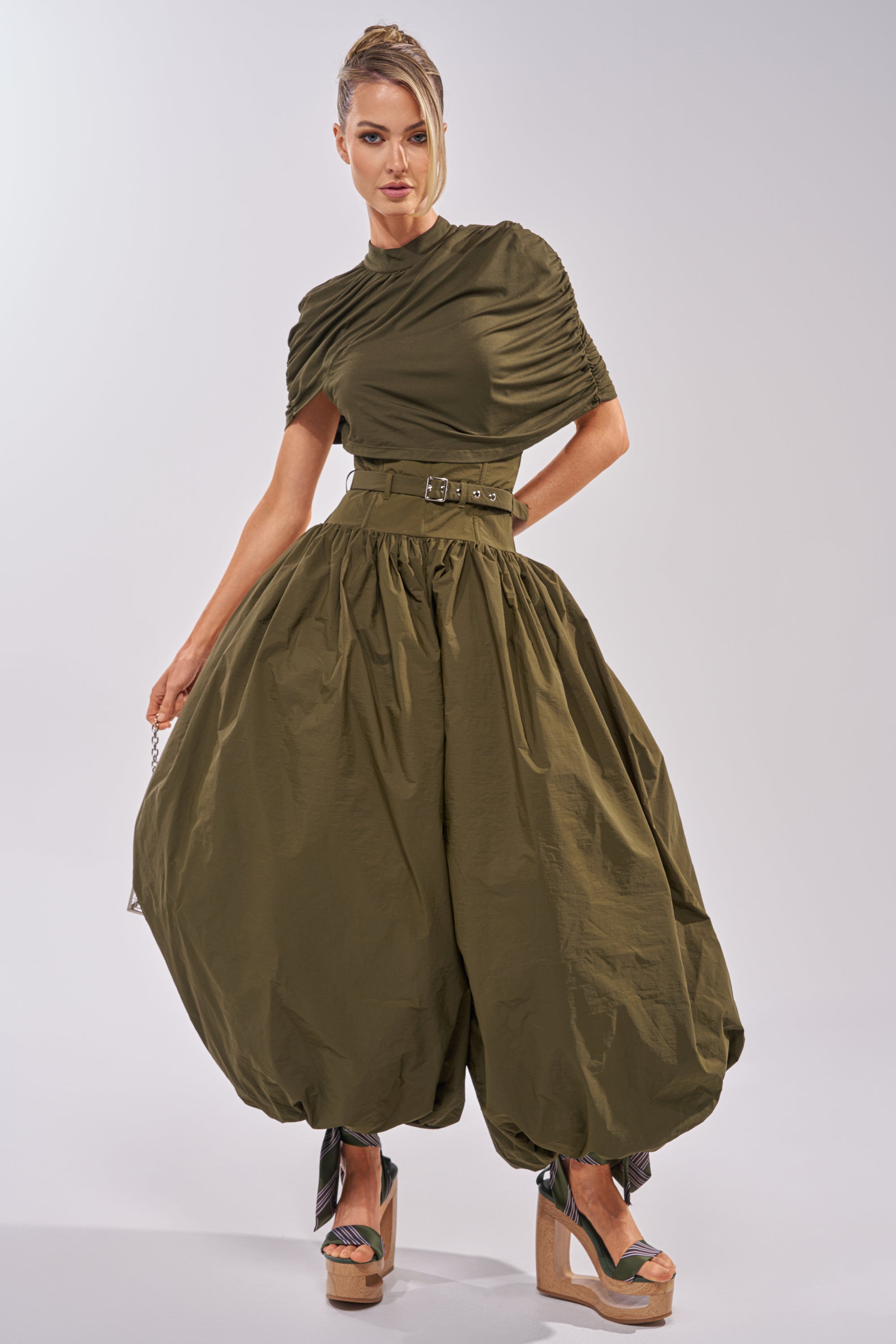 Wearing the SPIN ME AROUND POPLIN JUMPSUIT, which features an olive hue, a belted waist, and a voluminous balloon-style skirt, the woman pairs it with platform wedge sandals as she stands against a plain background.