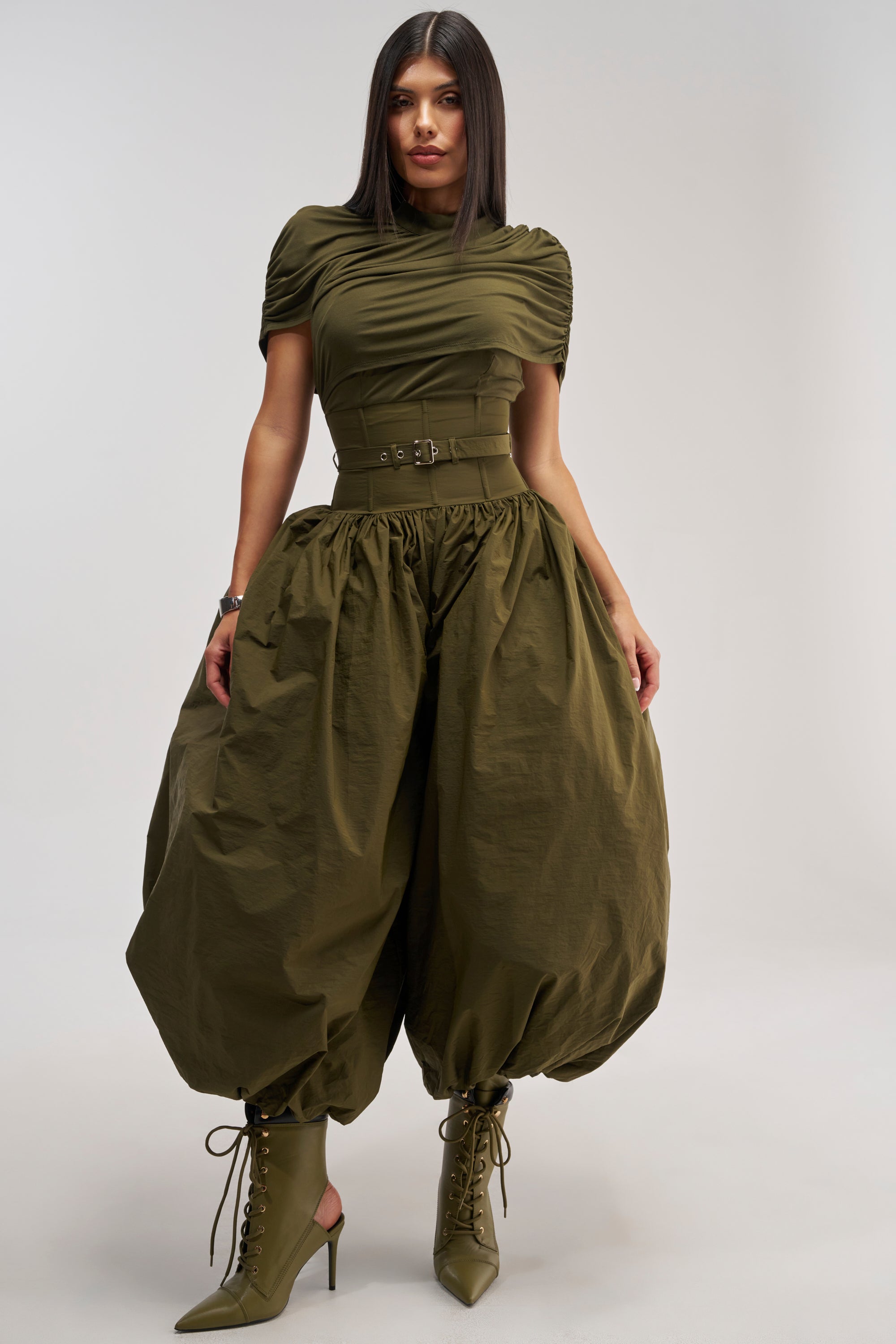 Wearing the "SPIN ME AROUND POPLIN JUMPSUIT" in olive green, which features a ruched top, wide belted waist, and dramatically voluminous balloon-shaped legs, a woman poses against a plain light background. Her outfit is completed with coordinating heeled lace-up boots.