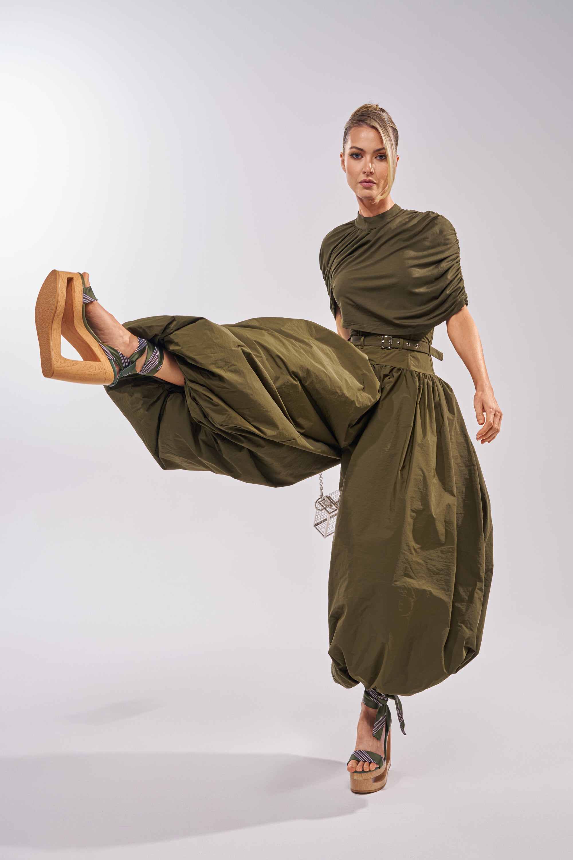 A woman in the SPIN ME AROUND POPLIN JUMPSUIT and platform sandals poses with one leg lifted, highlighting the jumpsuit's flowing fabric and dramatic shape against a plain light background. 1