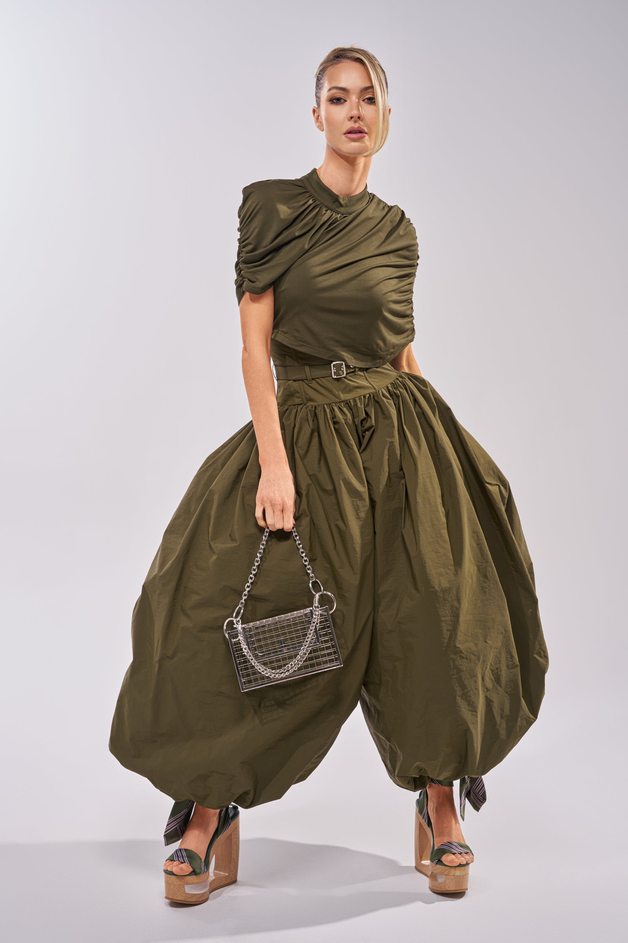 A woman models the SPIN ME AROUND POPLIN JUMPSUIT, featuring ruched details and balloon-style pants. She holds a small chain-strap handbag and wears platform sandals against a plain, light-colored background. 2