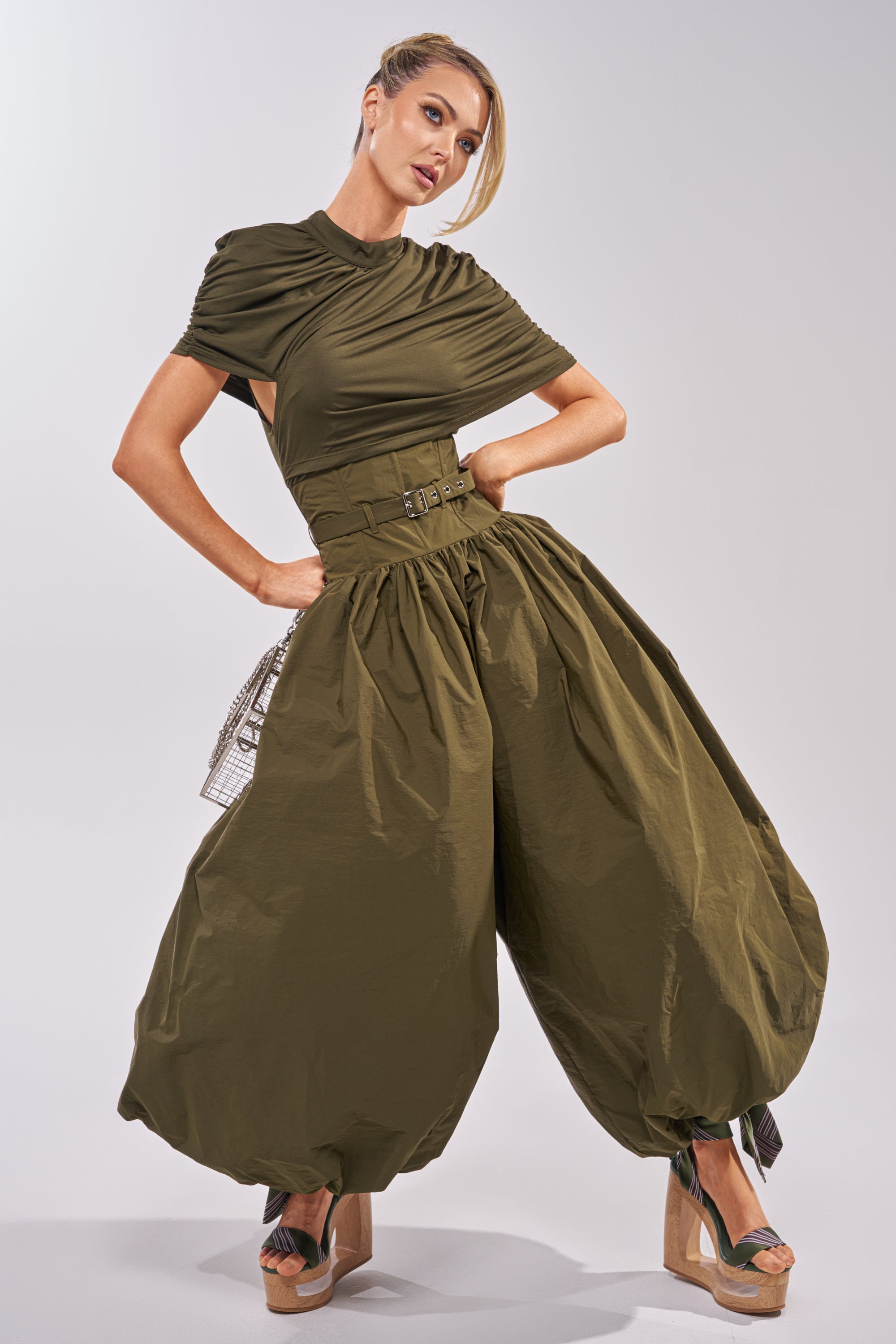 A woman poses in the SPIN ME AROUND POPLIN JUMPSUIT, showcasing its olive hue, draped fitted top, wide belted waist, and balloon-style pants gathered at the ankles. Platform sandals and an elegant updo complete her sculptural look. 3