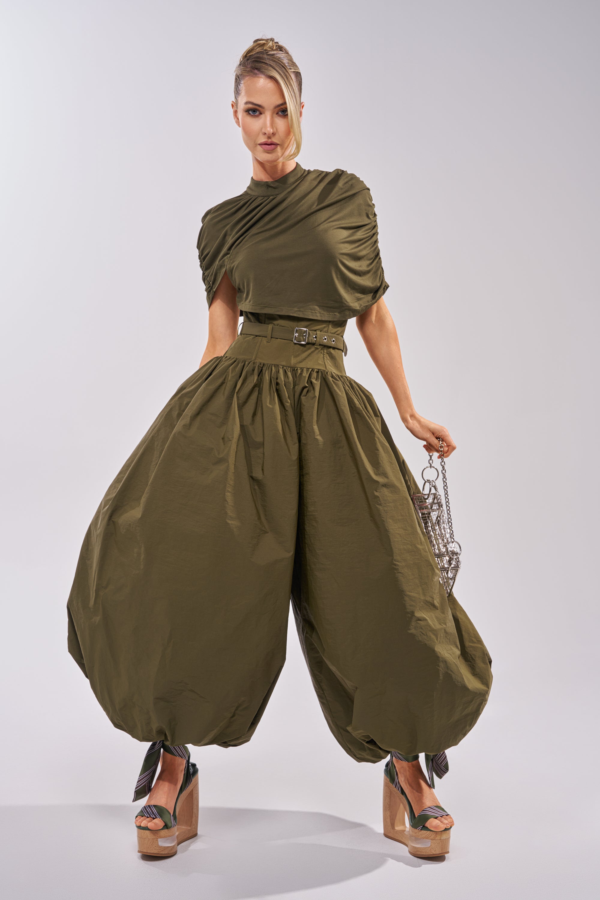 A woman models the SPIN ME AROUND POPLIN JUMPSUIT, featuring dramatic voluminous pants and a belted top. She completes the look with platform heels and a silver chain bag, standing against a plain white background. 4