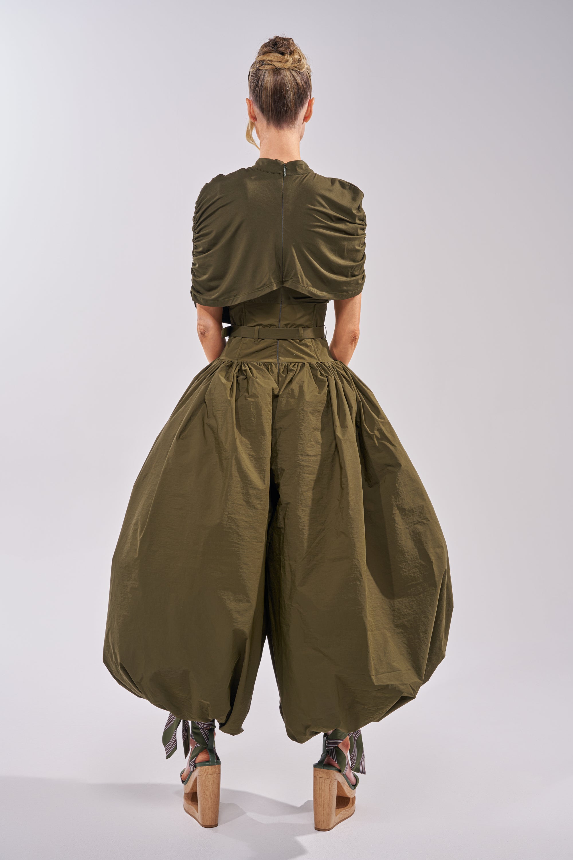 A person faces away wearing the SPIN ME AROUND POPLIN JUMPSUIT in olive with a ruched top, balloon legs, and corseted waist. Their hair is in a bun, paired with chunky high-heeled sandals with ribbon ties, set against a plain light background. 6