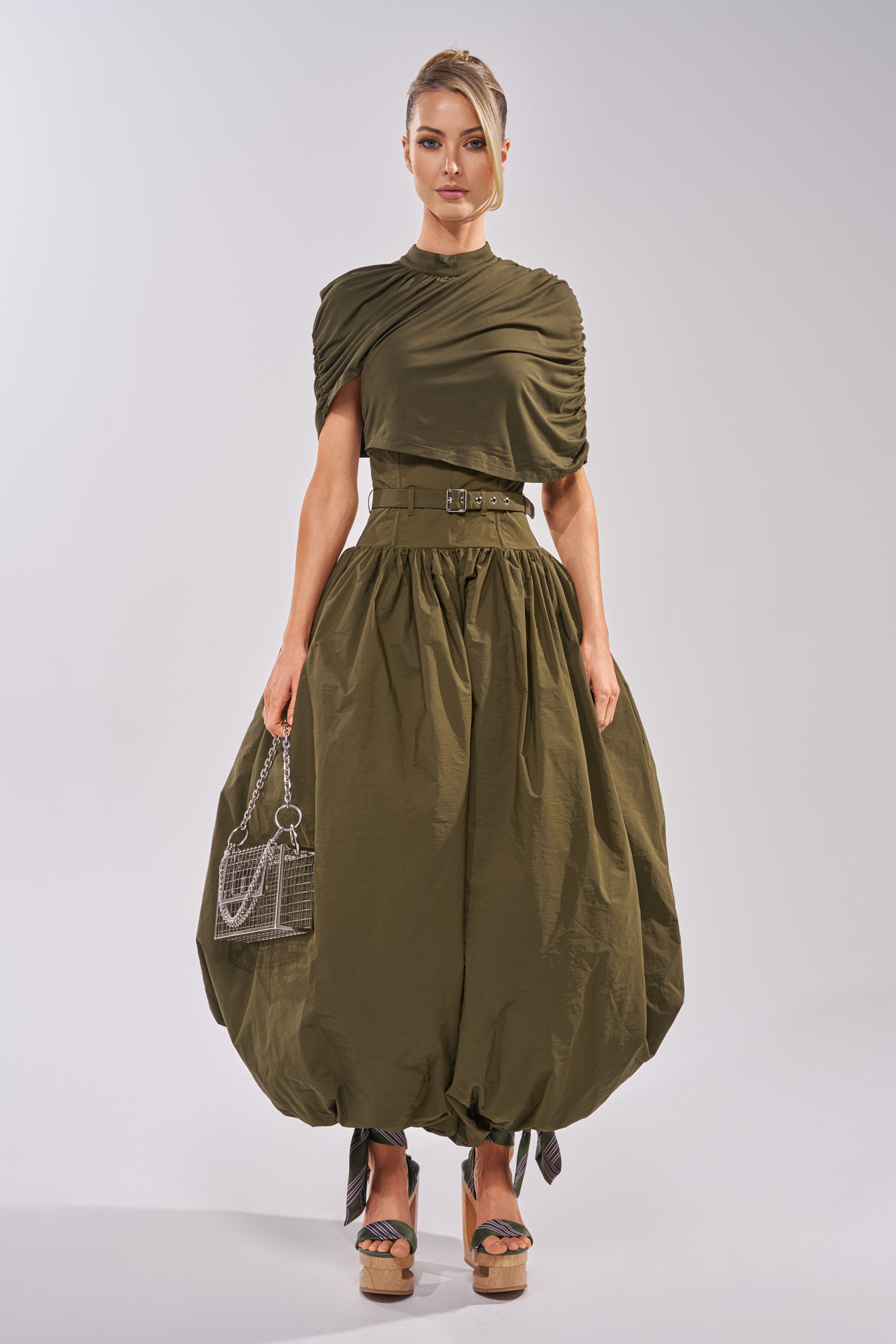 A woman stands against a plain background wearing the SPIN ME AROUND POPLIN JUMPSUIT with a ruched top, balloon legs, and a corseted waist. She carries a small transparent handbag and wears platform sandals. 7