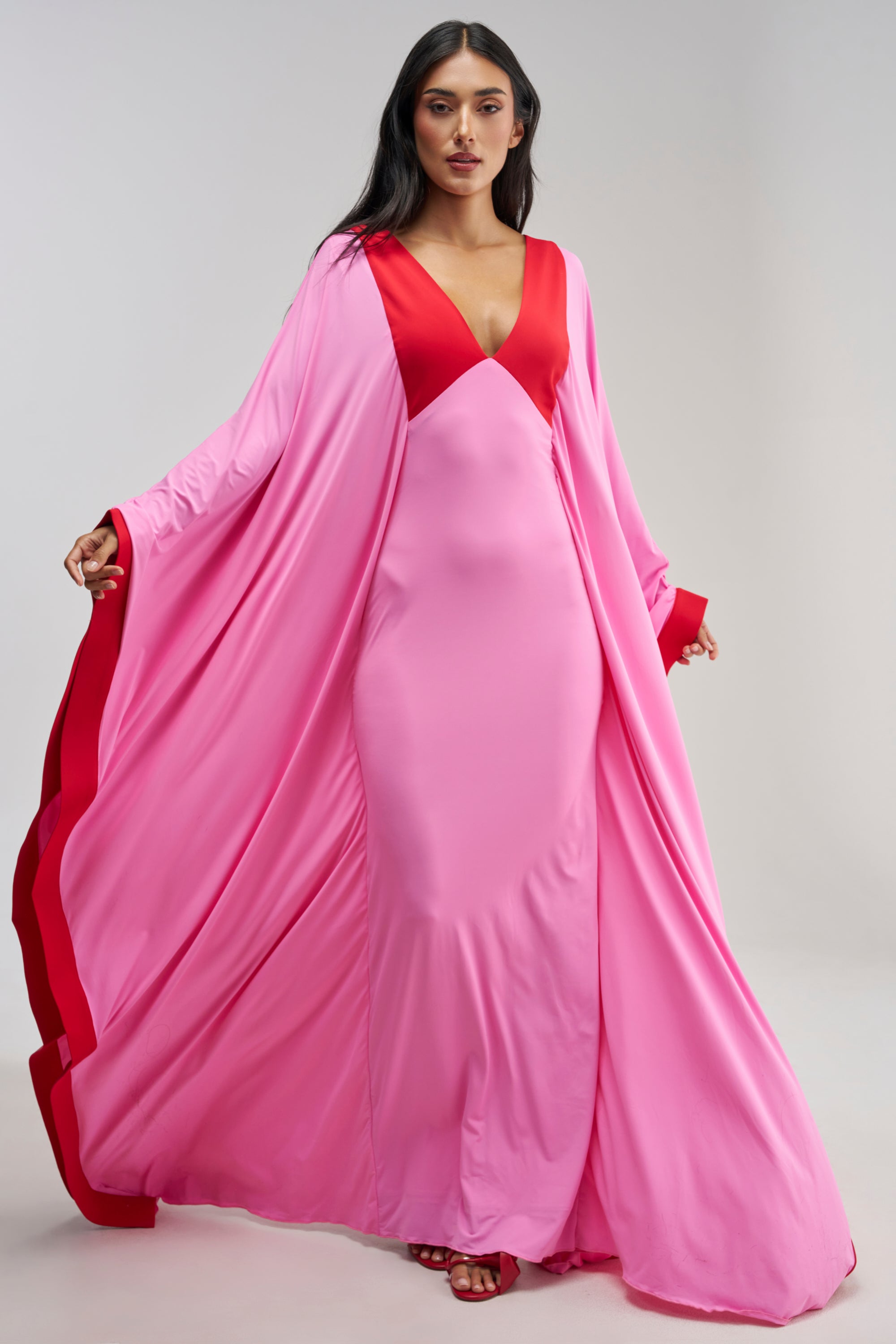 A woman models the GUEST OF HONOR MAXI DRESS IN PINK, a flowing, color-blocked maxi dress featuring striking red accents on the neckline, cuffs, and inner edges, standing against a plain light background.