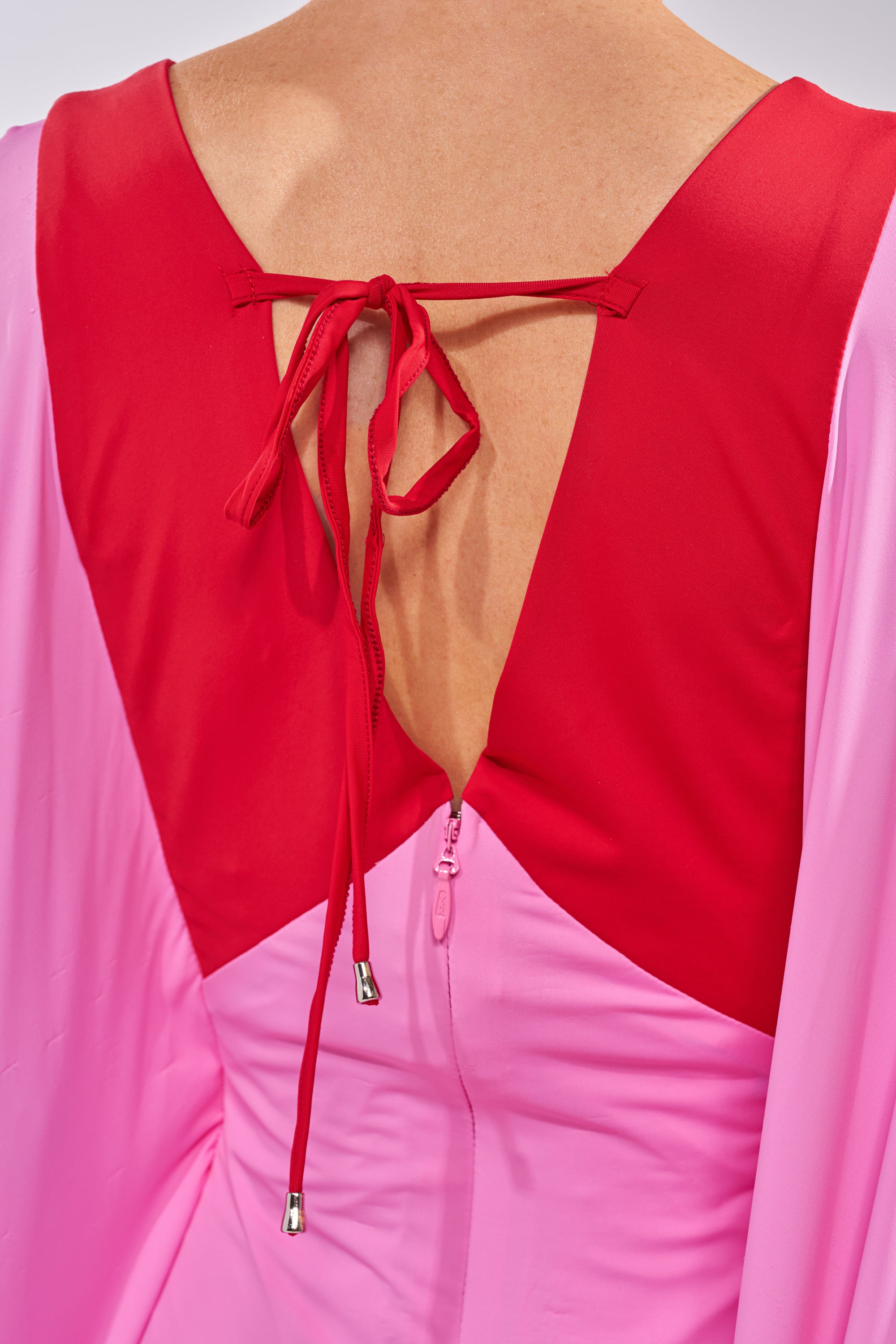 A close-up of a person’s back wearing the GUEST OF HONOR BODYCON MAXI DRESS IN PINK, featuring a deep red V-shaped neckline, back zipper, and matching red tie closure at the top. 5