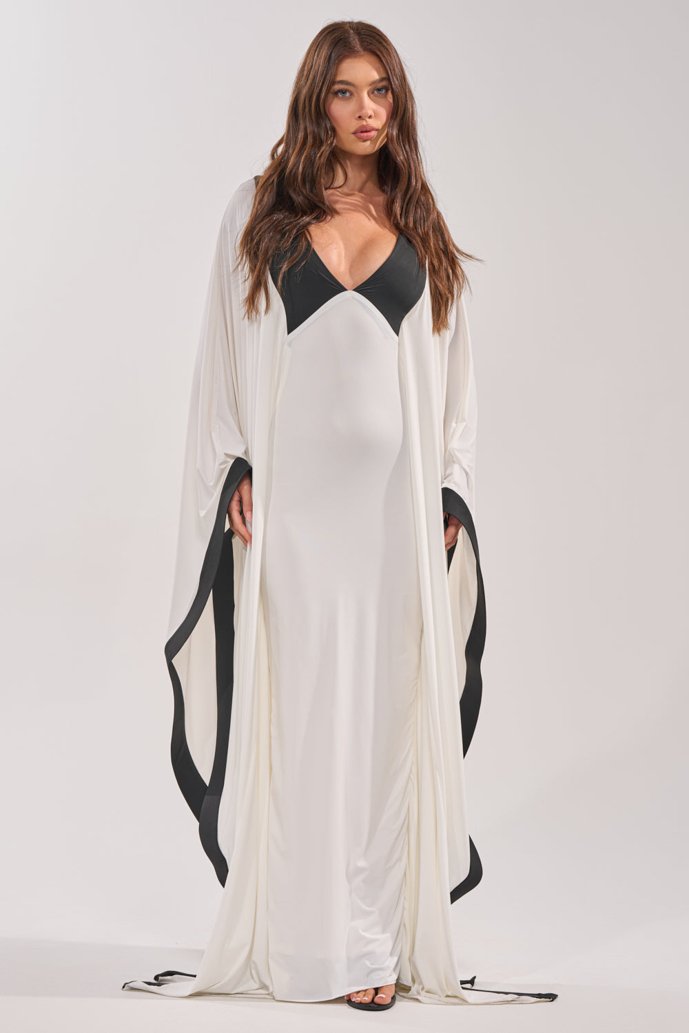 A woman poses facing forward in the GUEST OF HONOR BODYCON MAXI DRESS, featuring a monochrome design with black accents on the neckline and hem. Her loose, wavy hair contrasts with the plain, light background. 0