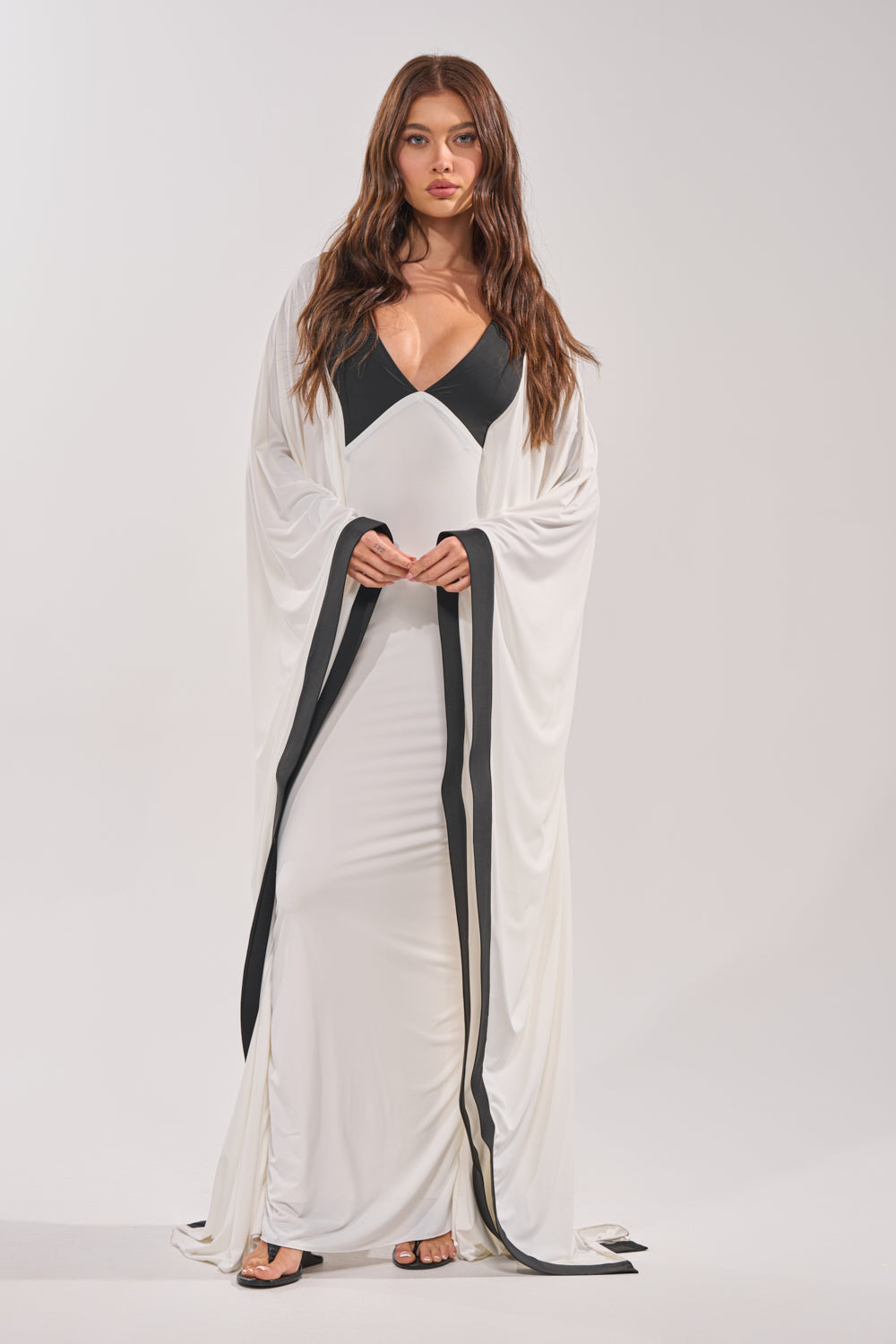 A woman with long brown hair wears the GUEST OF HONOR BODYCON MAXI DRESS in white with black trim and a deep neckline, paired with open-toe black heels. She stands facing forward against a plain light background, exuding monochrome glamour. 1