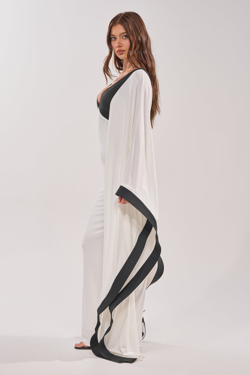 A woman wearing the GUEST OF HONOR BODYCON MAXI DRESS in white with black trim and a flowing cape-like sleeve stands against a plain white background, gazing at the camera with a neutral expression. 2