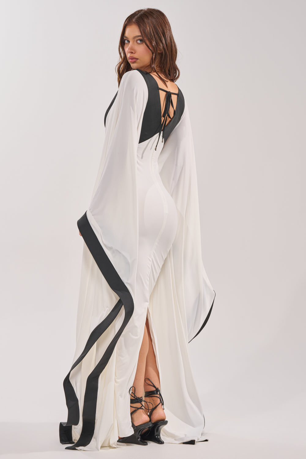 A woman poses in the GUEST OF HONOR BODYCON MAXI DRESS, featuring a long, fitted silhouette with flowing black-trimmed sleeves and a black back detail. She looks over her shoulder in strappy black heels against a plain light background. 3