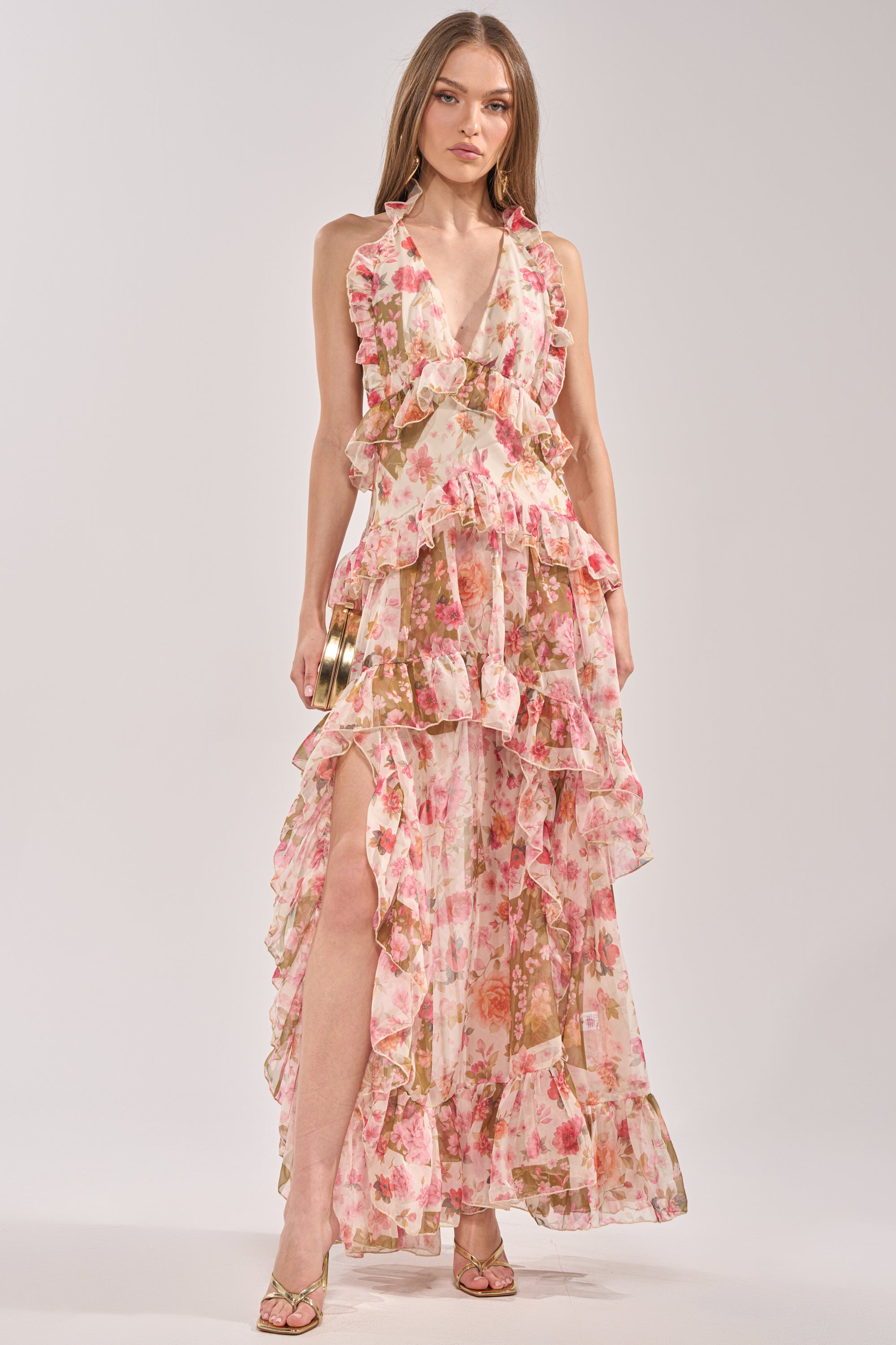 A woman faces the camera in the SUMMER STROLL FLORAL CHIFFON MAXI DRESS, a sleeveless pink floral maxi with ruffles, deep V-neckline, and high slit—ideal for special occasions and styled with strappy heels.