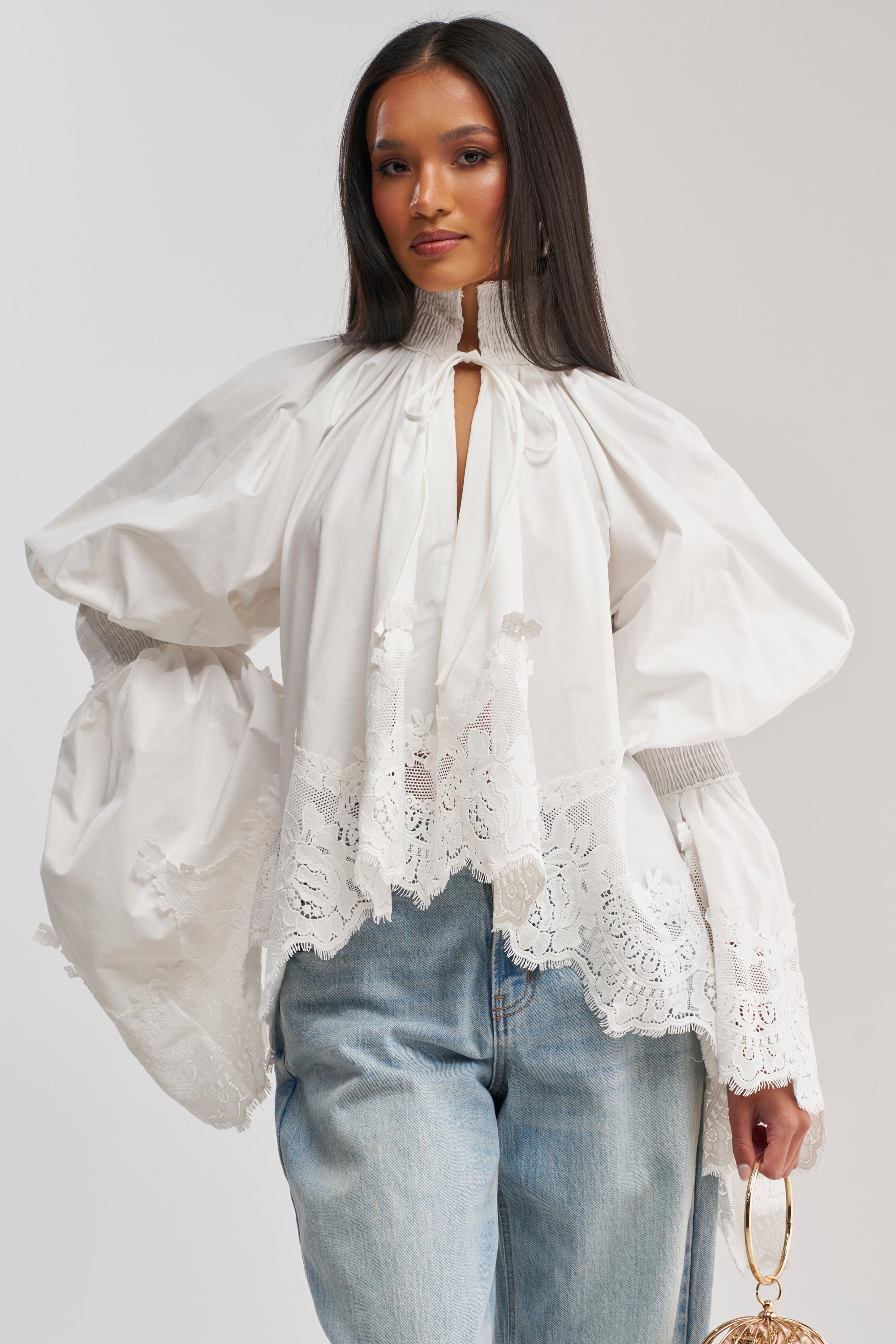A woman wears the MARIA LACE TRIM POPLIN BLOUSE with billowy sleeves and lace accents, paired with light blue jeans. She holds a small round handbag and stands against a simple light background.