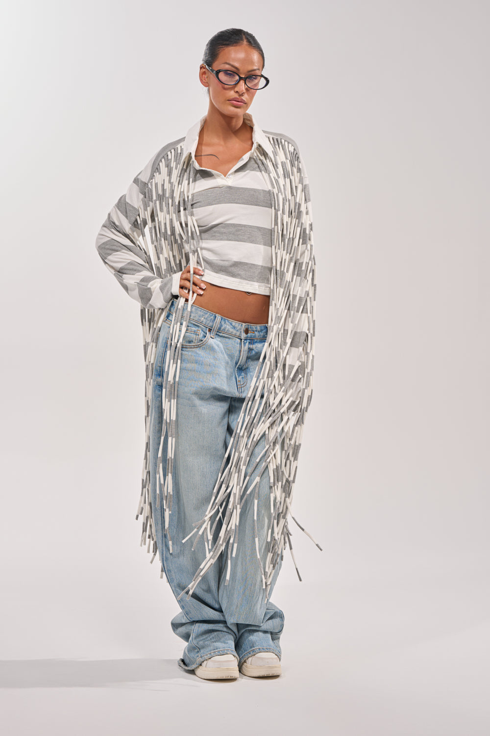 A woman in glasses and the SOMETHING LIGHT FRINGE TOP, paired with a long fringe cardigan, loose blue jeans, and white sneakers, stands confidently against a plain white background. 4
