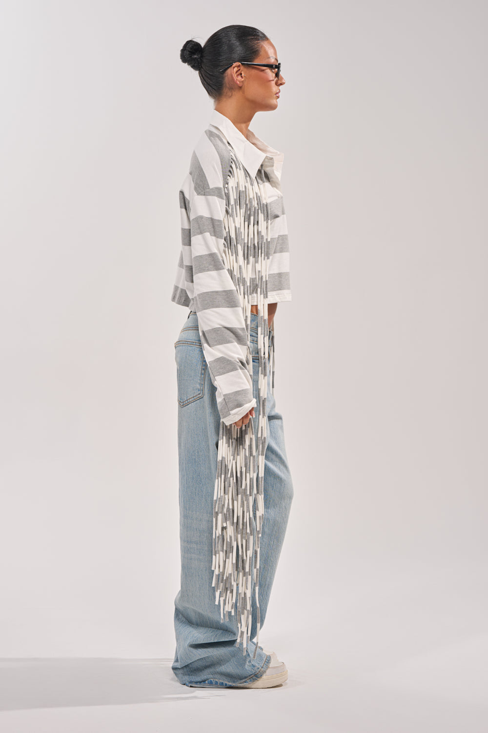 A woman with dark hair in a bun, sunglasses, loose light blue jeans, and white sneakers stands in profile against a plain light background wearing the SOMETHING LIGHT FRINGE TOP. 5