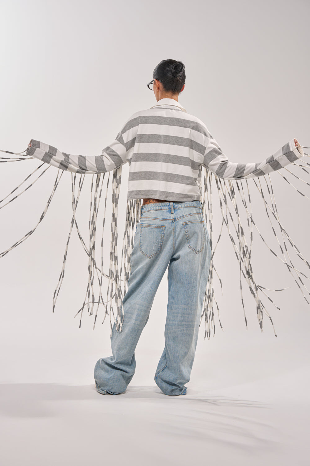 A person faces away with outstretched arms, wearing loose jeans and the SOMETHING LIGHT FRINGE TOP—a striped button-up featuring long, trailing fringe on the sleeves—against a plain, light background. 7