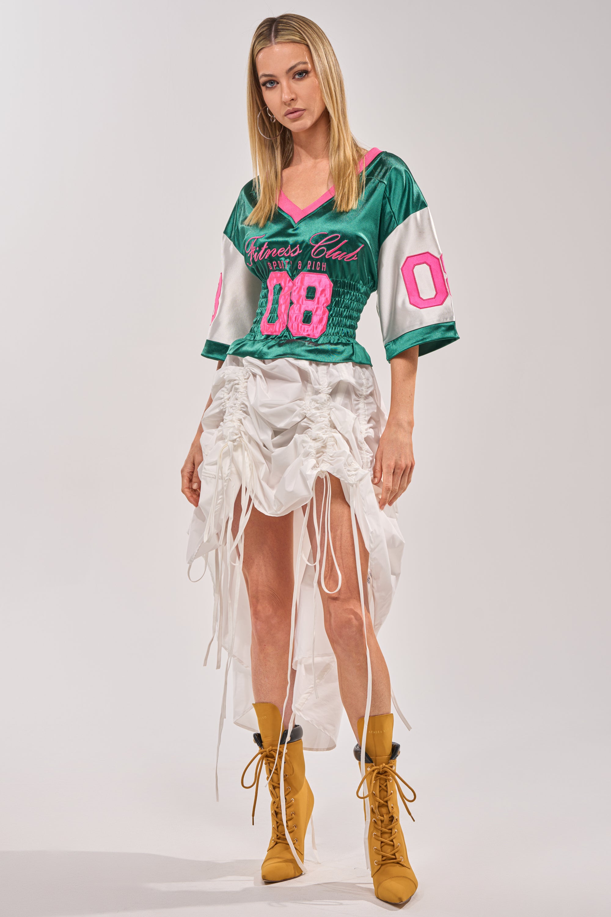 A woman wears the FIX YOUR GAME JERSEY MAXI DRESS in green and pink with "08" on the sleeves, styled with a white ruched high-low skirt and mustard yellow lace-up boots against a plain background. 0