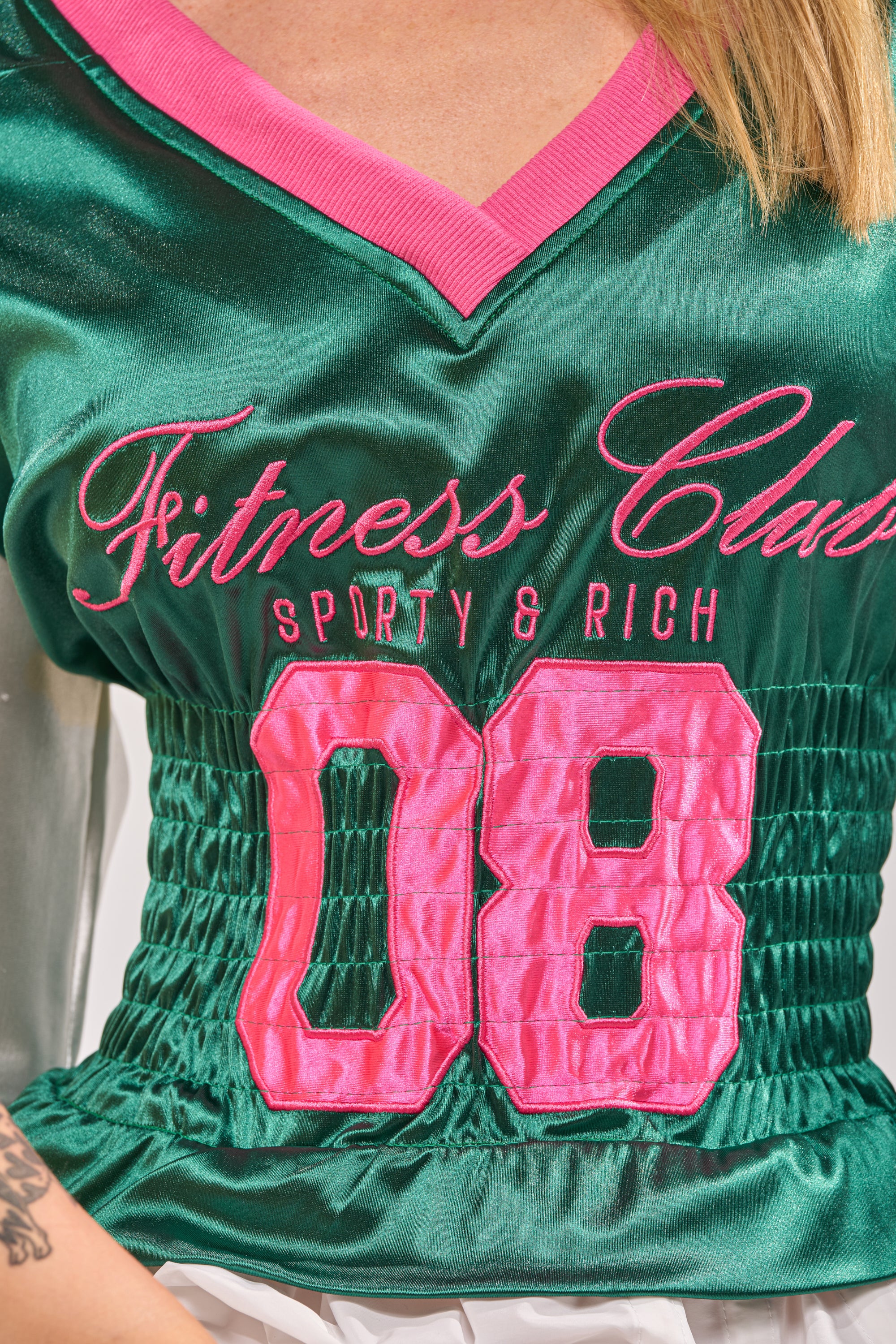 A person wears the FIX YOUR GAME JERSEY MAXI DRESS—a shiny green and pink varsity-inspired top with “Fitness Club Sporty & Rich 08” in pink lettering on the front. Only the torso and part of one arm are visible. 9