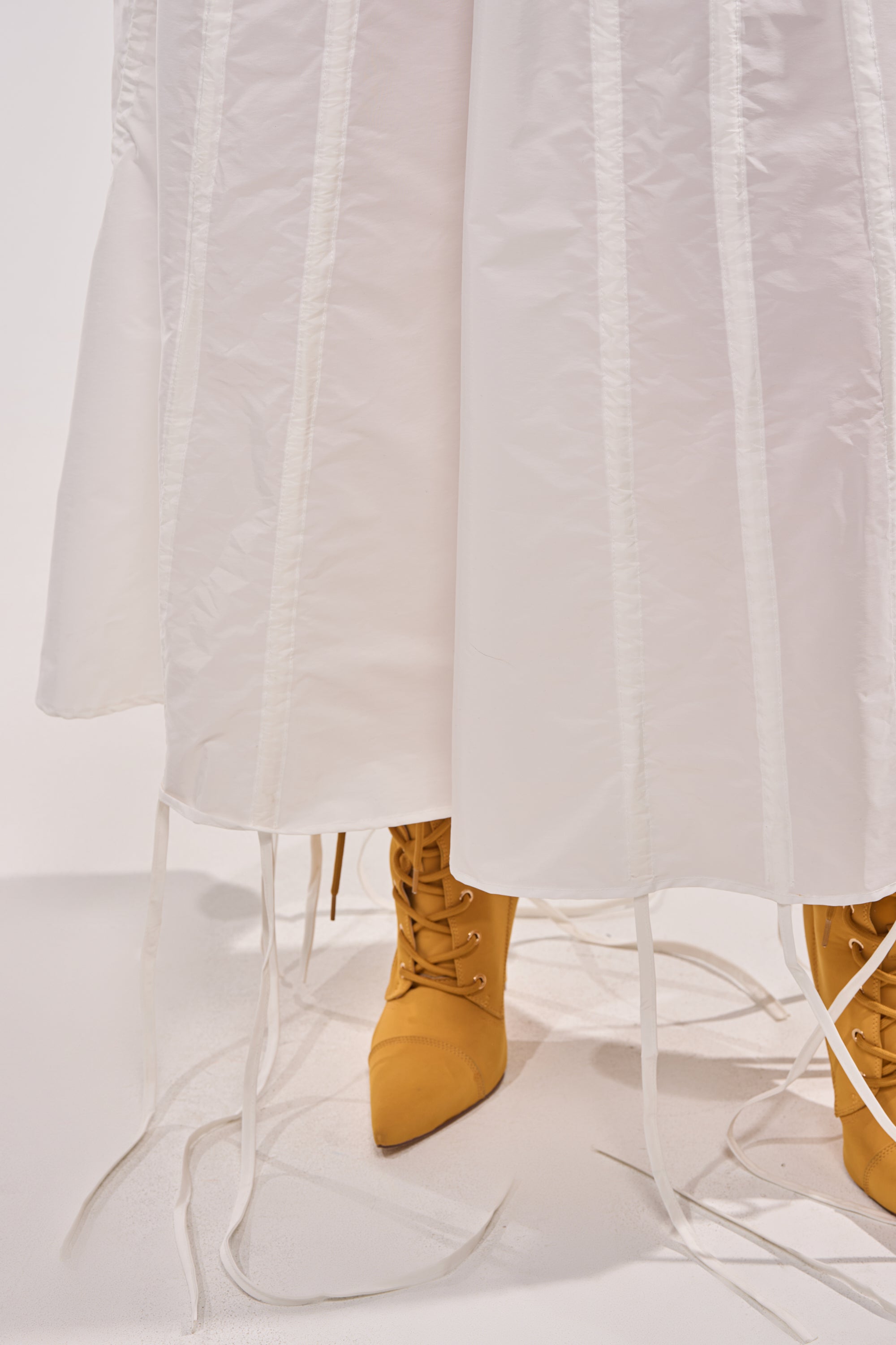 A person stands on a white floor wearing the FIX YOUR GAME JERSEY MAXI DRESS—a long, white dress with loose, dangling ribbons—paired with mustard yellow lace-up pointed boots for a sporty look. 12