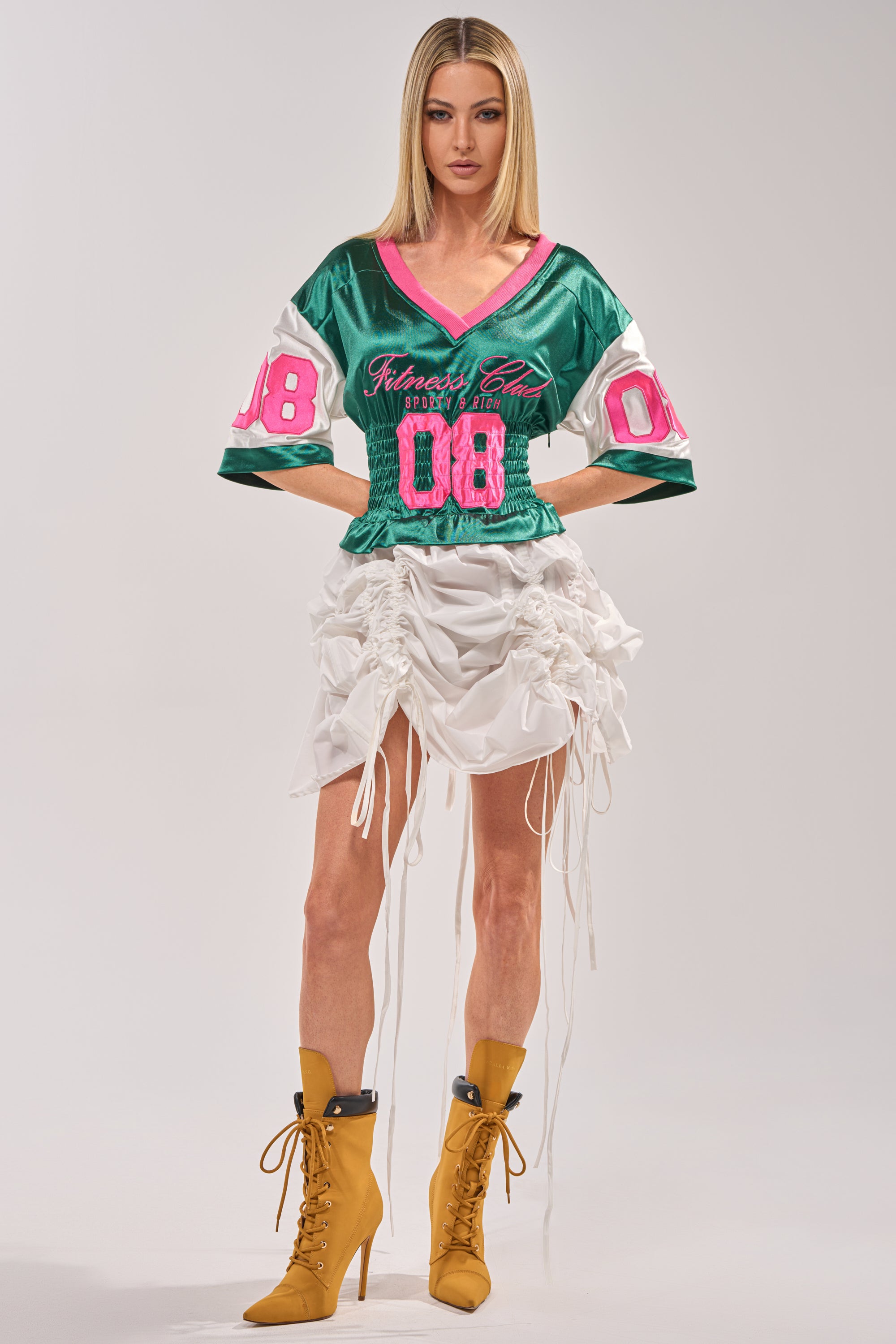 A woman with straight blonde hair faces forward in the FIX YOUR GAME JERSEY MAXI DRESS, a varsity-inspired green football jersey dress with pink details, against a plain background. She wears tan heeled boots and has a neutral expression. 1