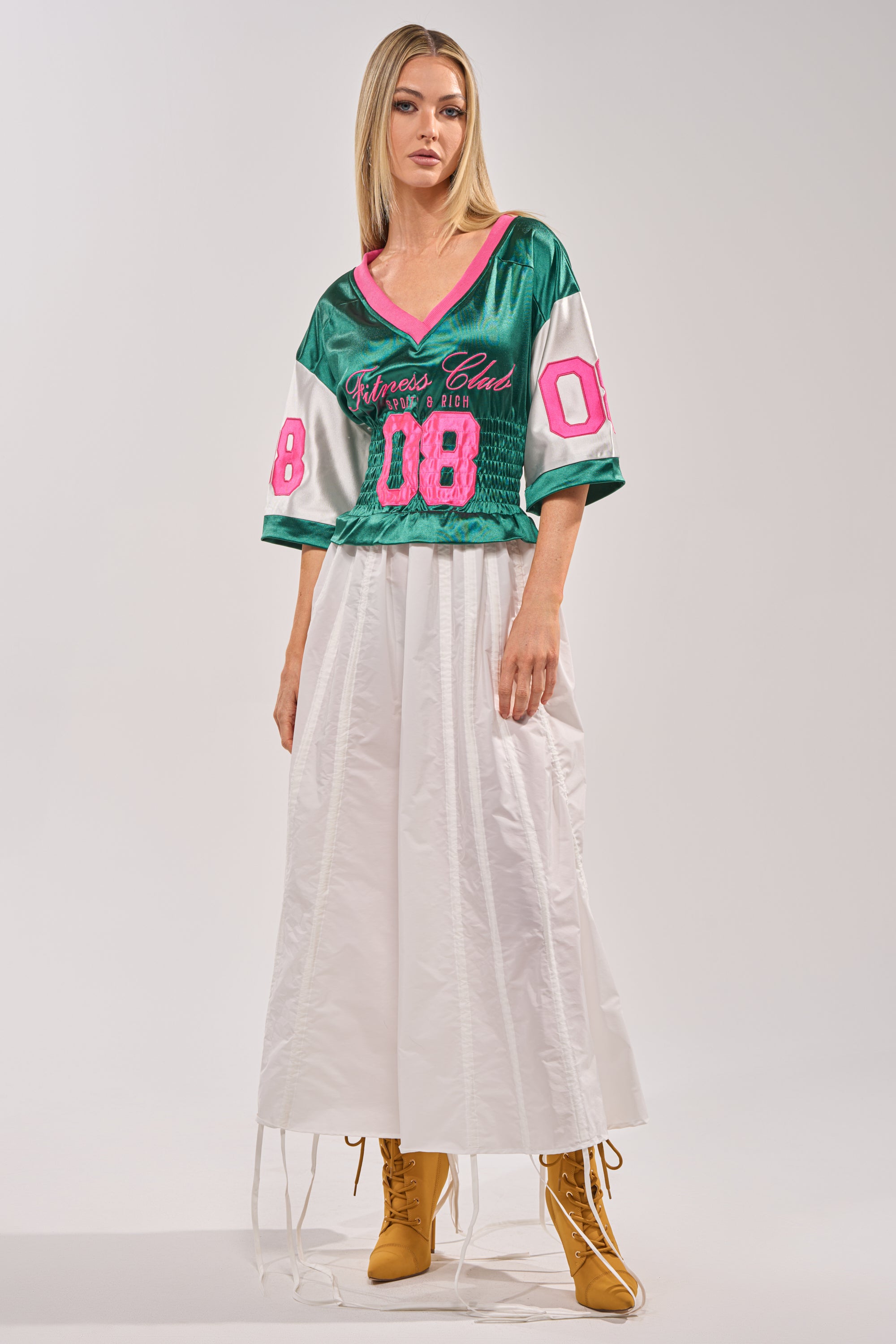 A woman with straight blonde hair and a neutral expression wears the FIX YOUR GAME JERSEY MAXI DRESS, a varsity-inspired green and pink piece with number 08, styled with tan lace-up boots. 2
