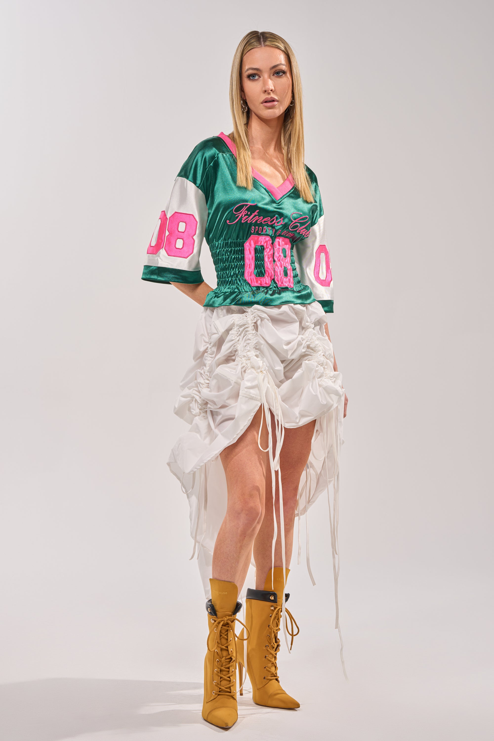 A woman poses against a light background in the FIX YOUR GAME JERSEY MAXI DRESS—a green varsity-style dress with pink "08" numbers—paired with mustard yellow lace-up boots. 4