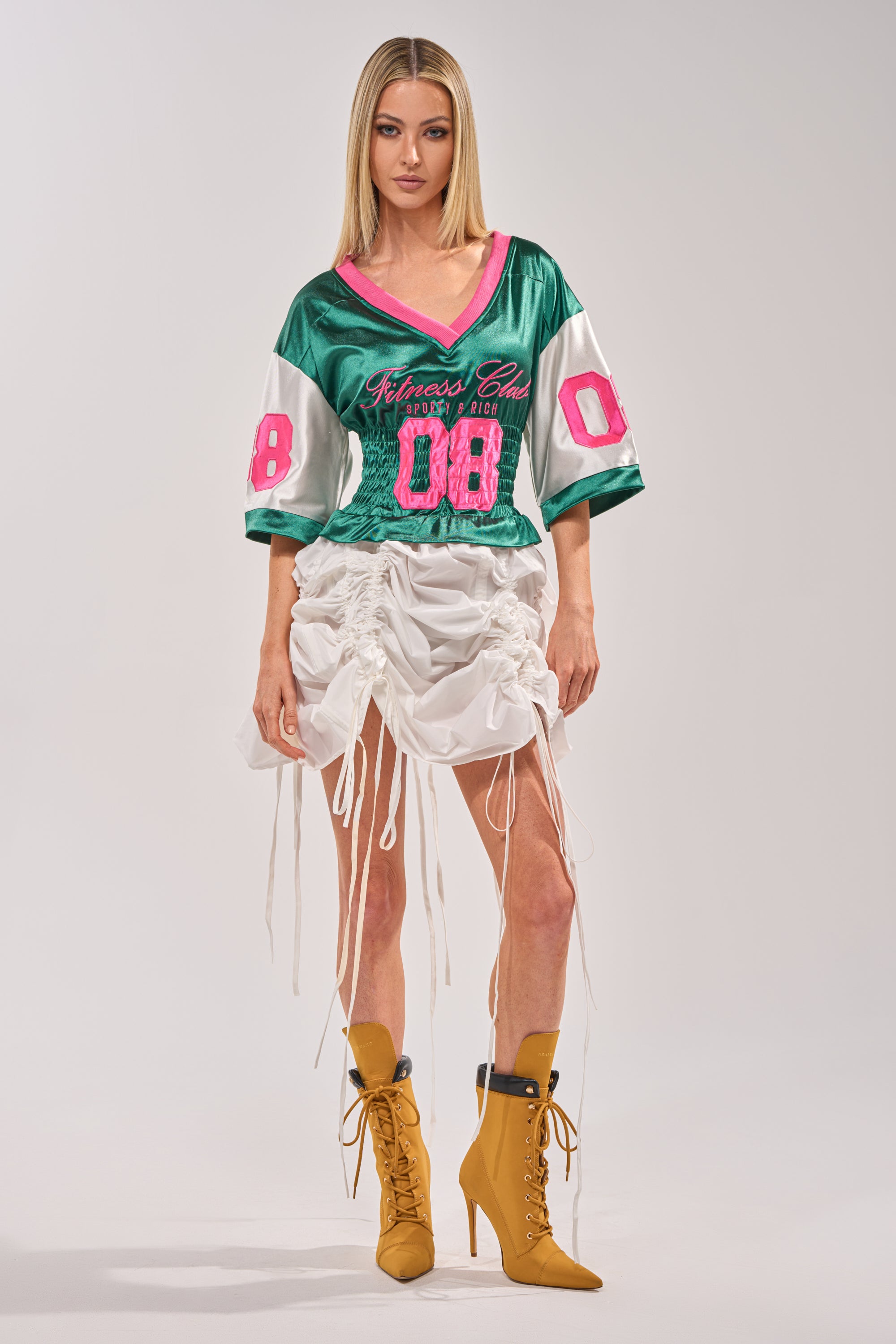 A woman with straight blonde hair and a neutral expression wears the FIX YOUR GAME JERSEY MAXI DRESS, a varsity-inspired piece with green and pink accents, paired with a white ruched mini skirt and mustard yellow lace-up ankle boots. 5