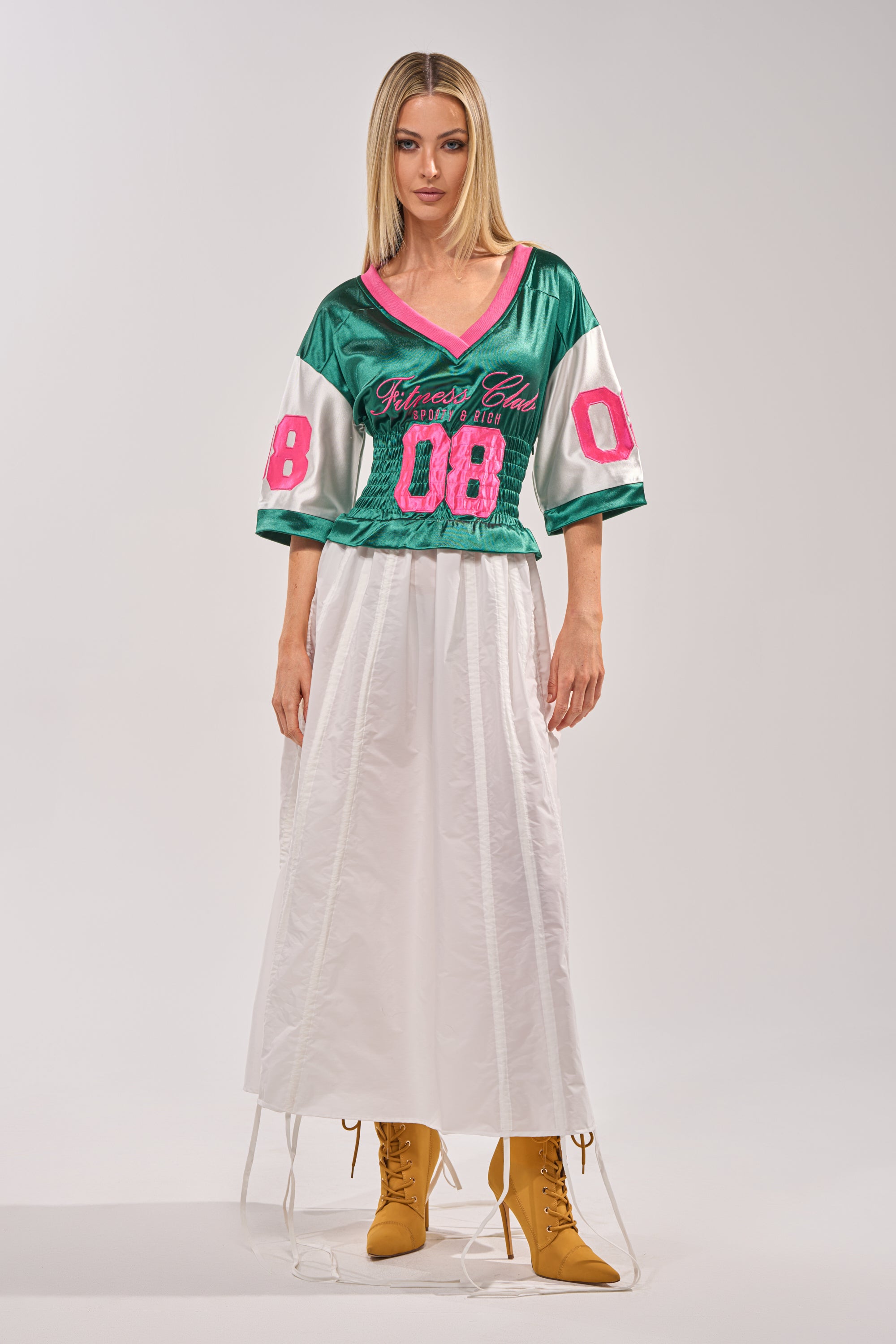 A woman with straight blonde hair and a neutral expression wears the FIX YOUR GAME JERSEY MAXI DRESS—green with pink "08" details—paired with a long white skirt and tan lace-up boots, standing against a light background. 6