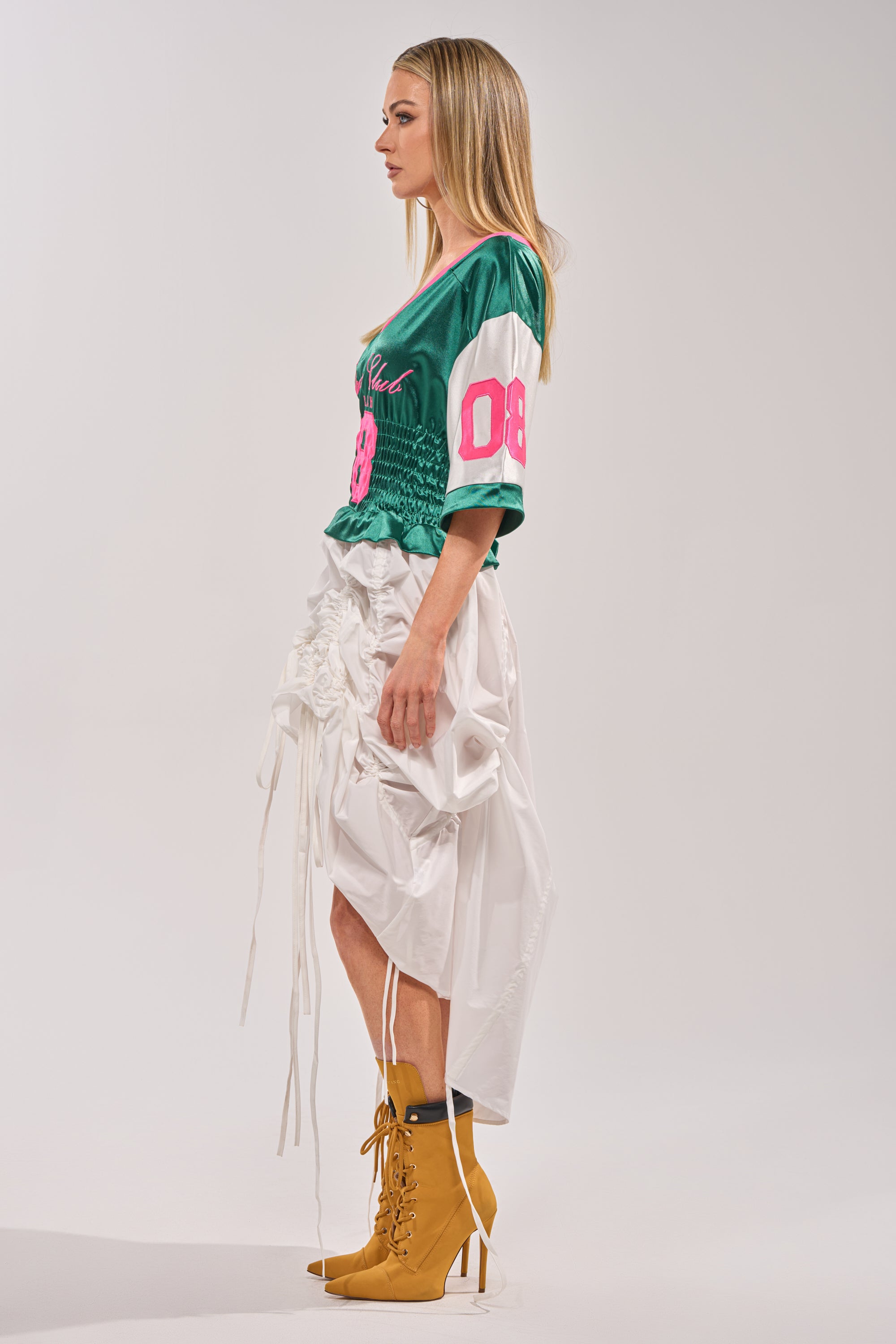 A woman stands in profile wearing the FIX YOUR GAME JERSEY MAXI DRESS—a green sporty chic dress with pink "08" lettering, styled with a white ruched skirt and tan lace-up heeled boots against a plain light background. 7