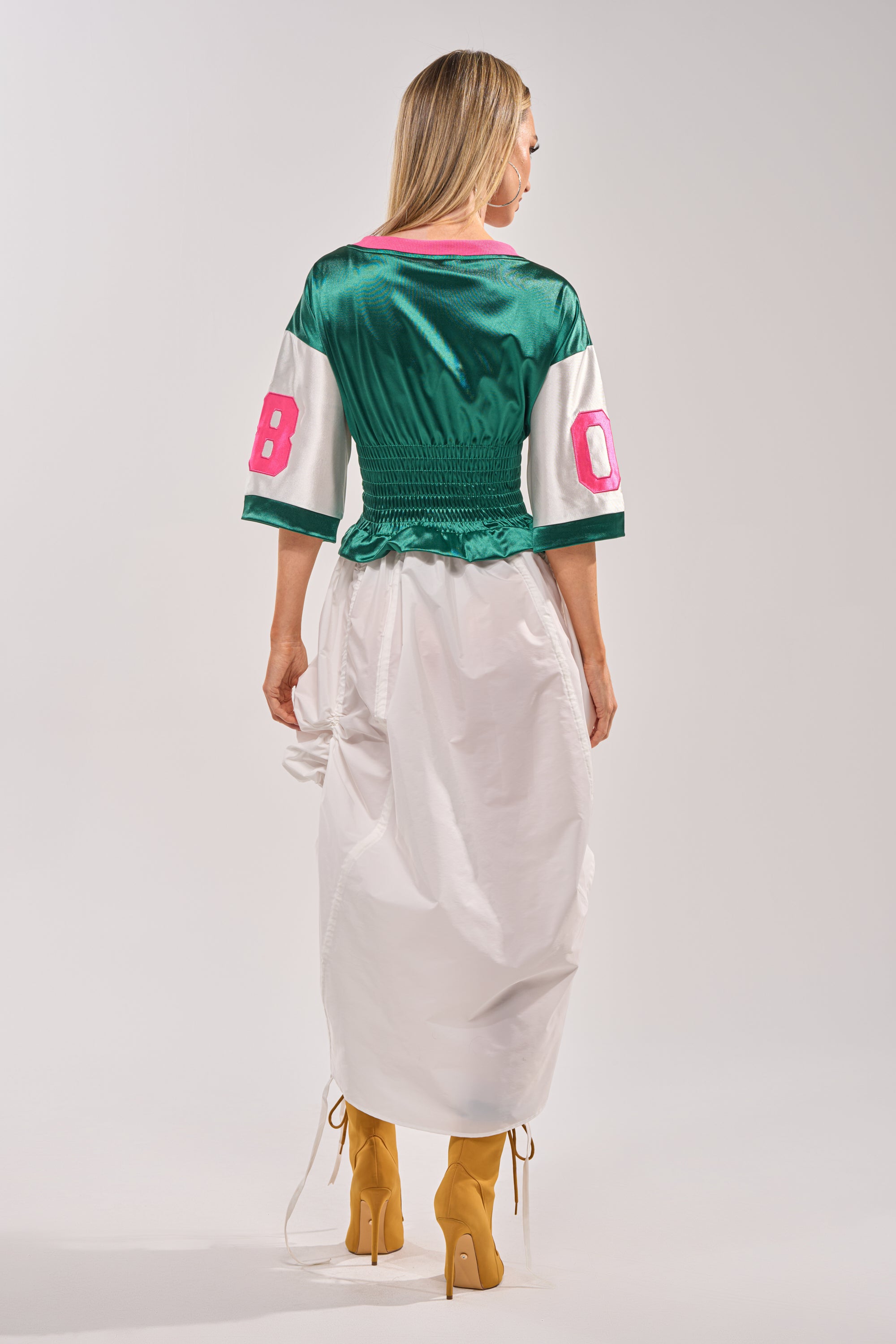 A woman faces away, wearing the FIX YOUR GAME JERSEY MAXI DRESS—green and white, varsity-inspired with pink sleeve numbers—styled with a long white skirt and yellow high-heeled boots against a plain background. 8