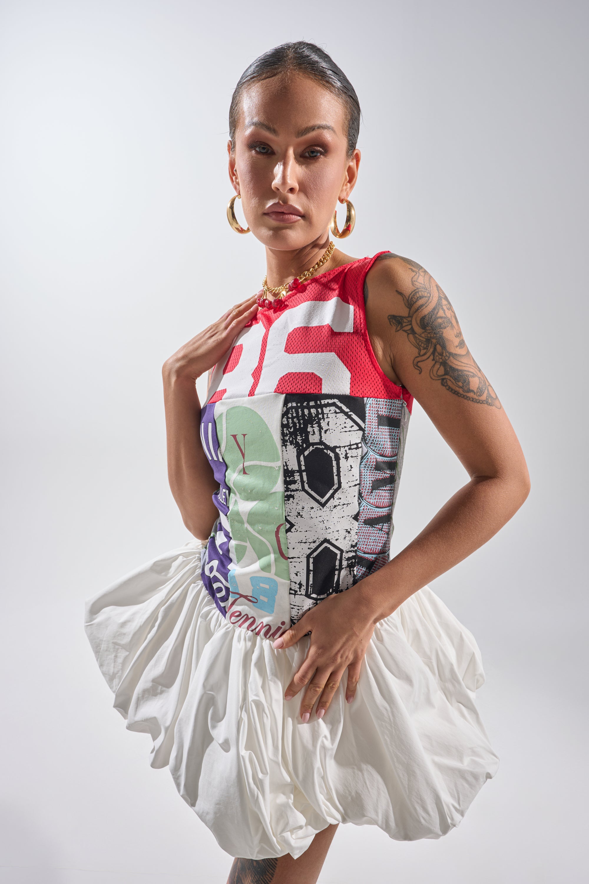 A person showcases the TEAM SPIRIT BUBBLE MINI DRESS, a sporty-chic style with bold red, white, and black patterns and a white skirt, accessorized with gold hoops and necklace, displaying arm tattoos against a plain background. 1