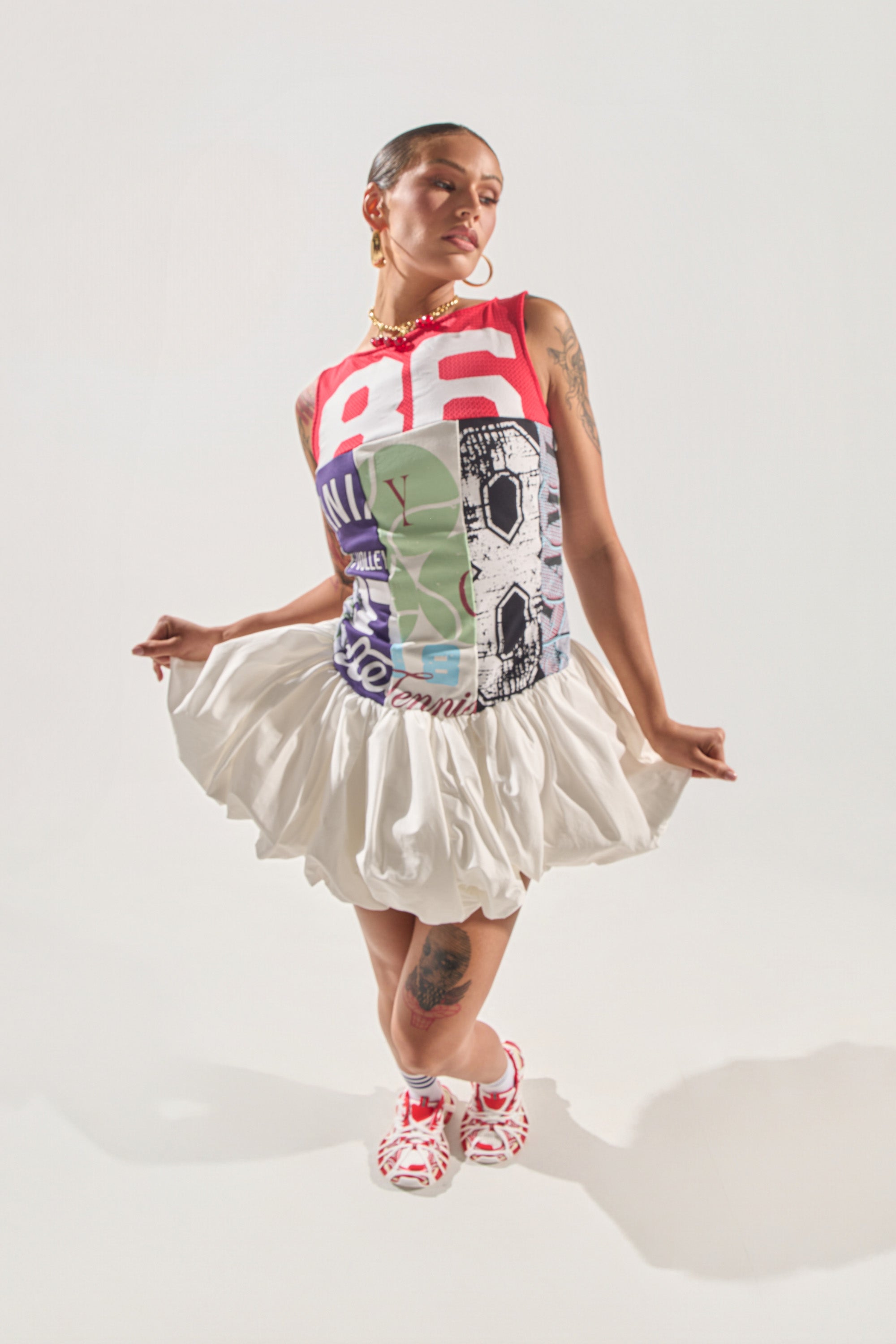 A woman models the TEAM SPIRIT BUBBLE MINI DRESS, featuring a sporty jersey style with the number 86, paired with a white ruffled skirt, red and white sneakers, gold jewelry, and hoop earrings against a plain white background. 2