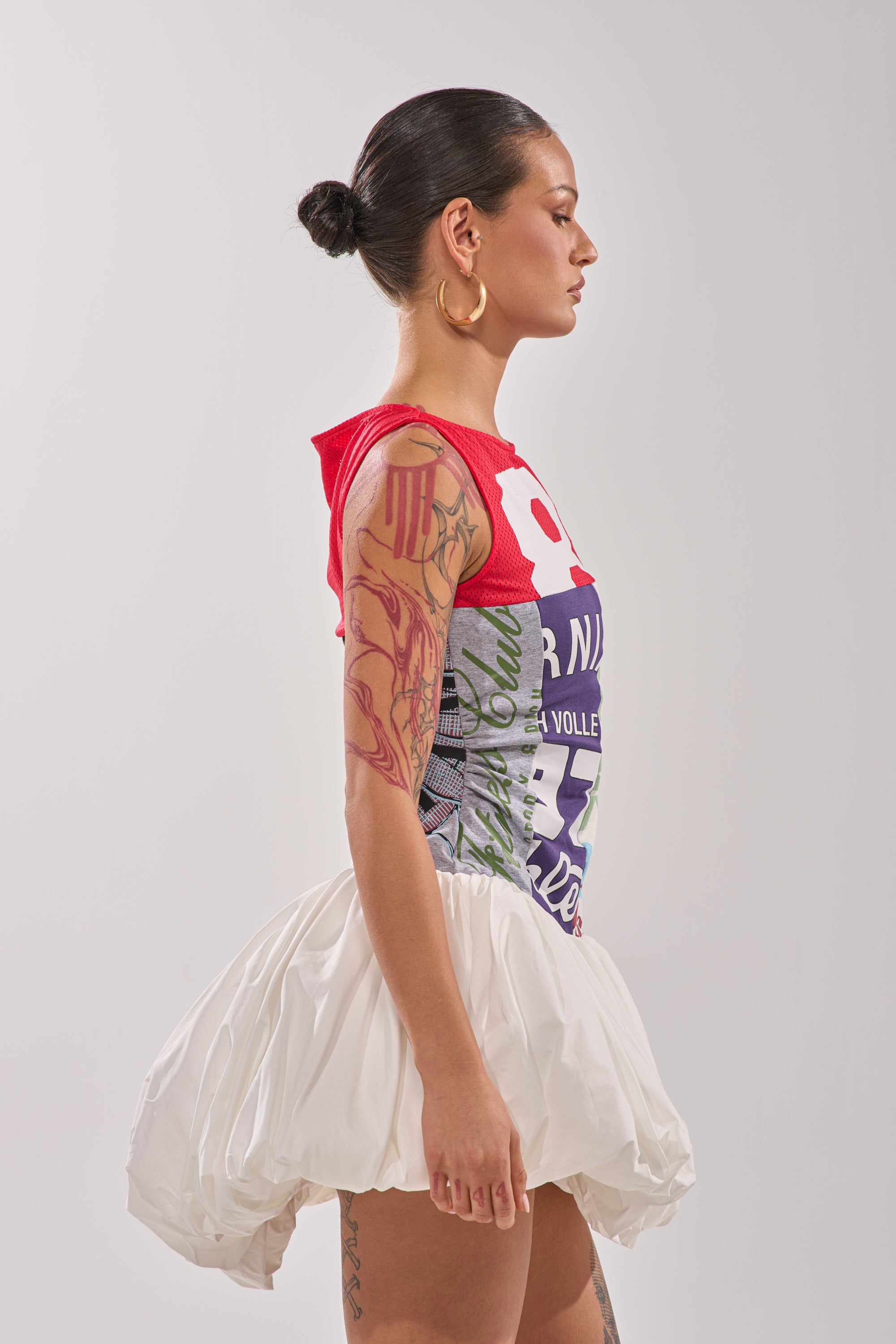 A woman with tied-back hair and arm tattoos stands in profile, modeling the sporty-chic TEAM SPIRIT BUBBLE MINI DRESS in red and multicolored tones against a plain, light background. 6