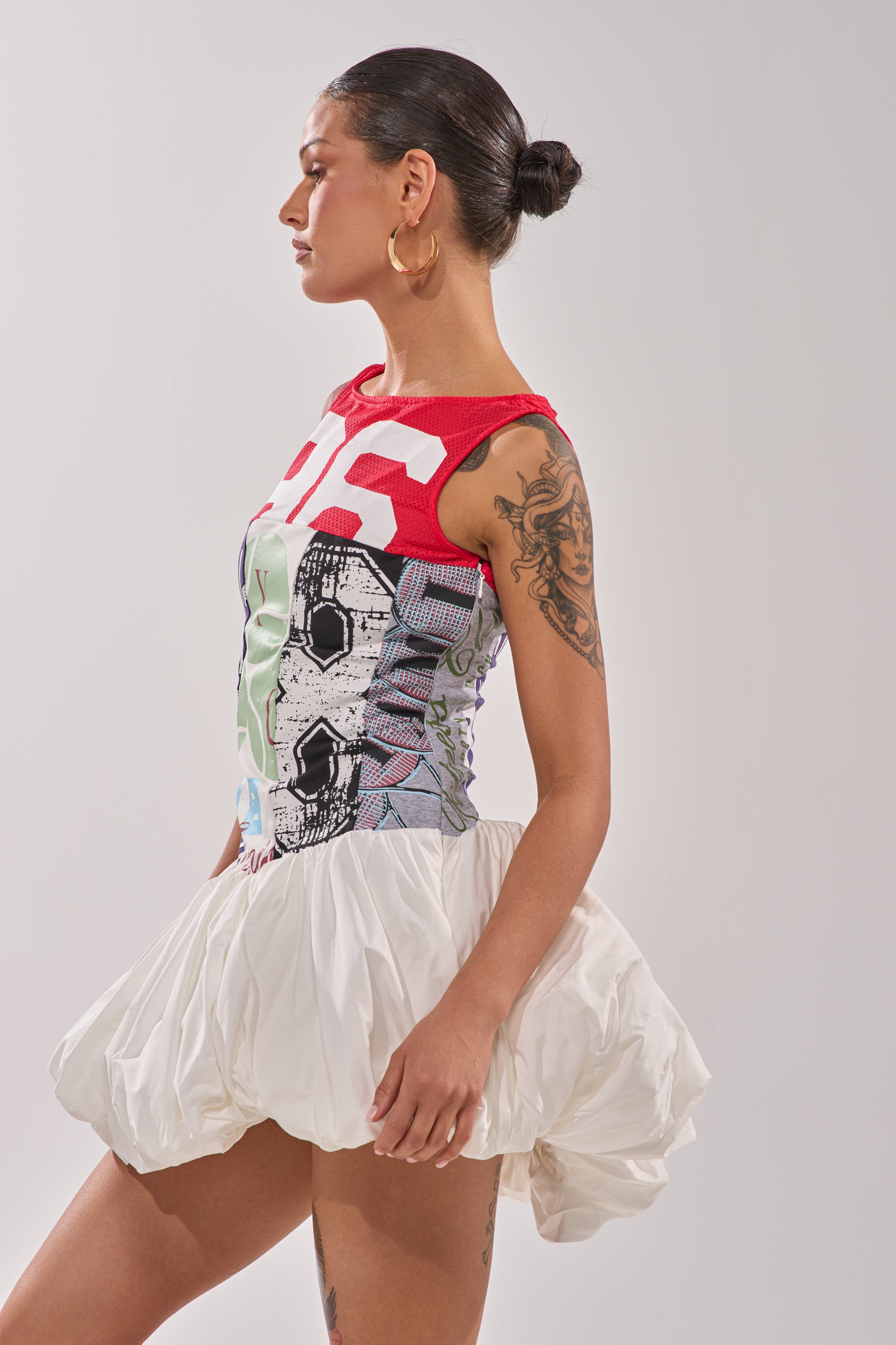 A woman with a tattoo on her arm models the TEAM SPIRIT BUBBLE MINI DRESS, featuring a red graphic print top and a short, white puffy skirt. She stands in profile against a plain light background. 7