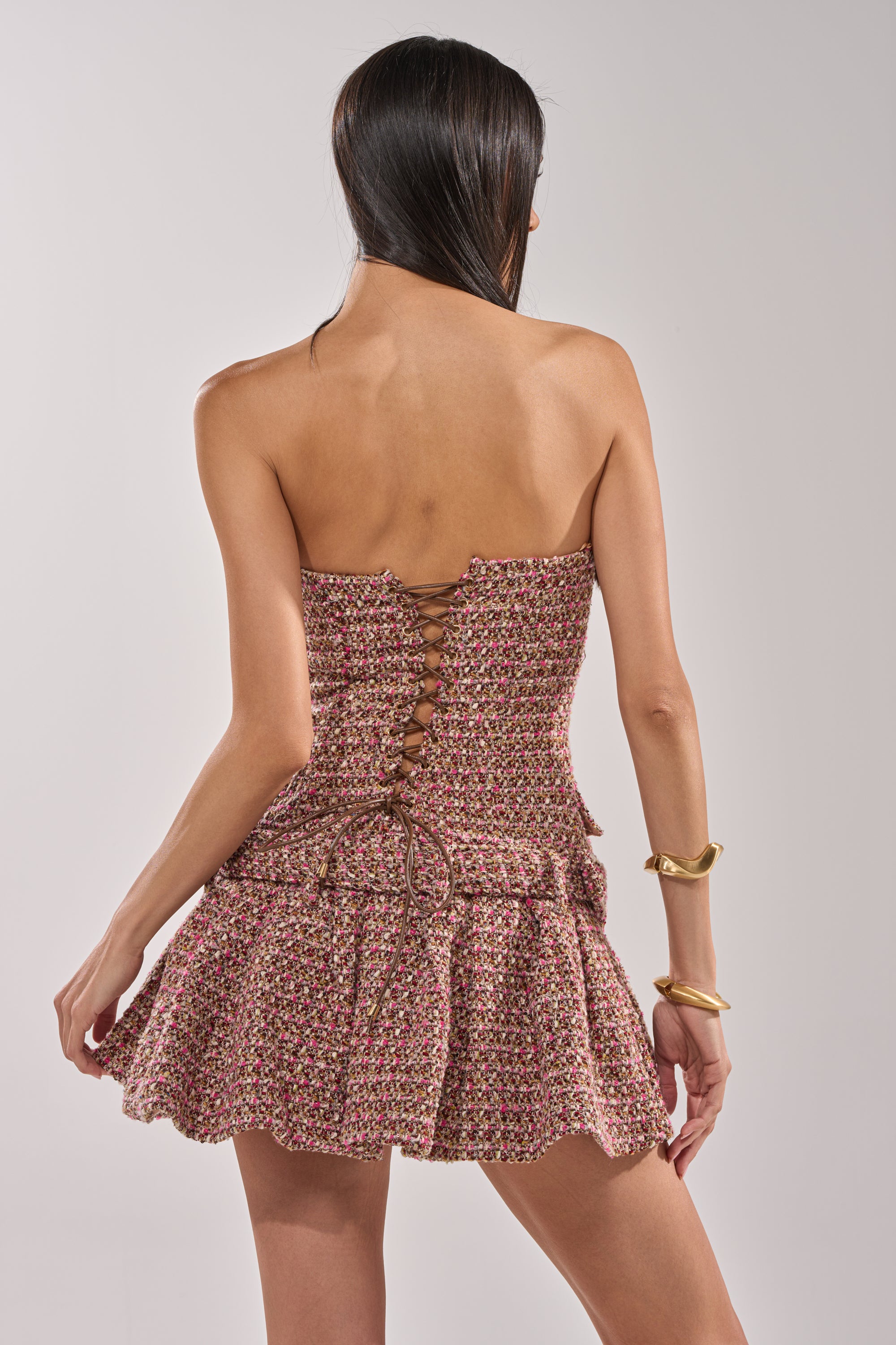 A woman with long dark hair, seen from the back, wears the SWEET CAROLINE MINI TWEED DRESS—a strapless mini with a ruffled skirt and lace-up back—paired with two chunky gold bracelets on her right wrist. 3