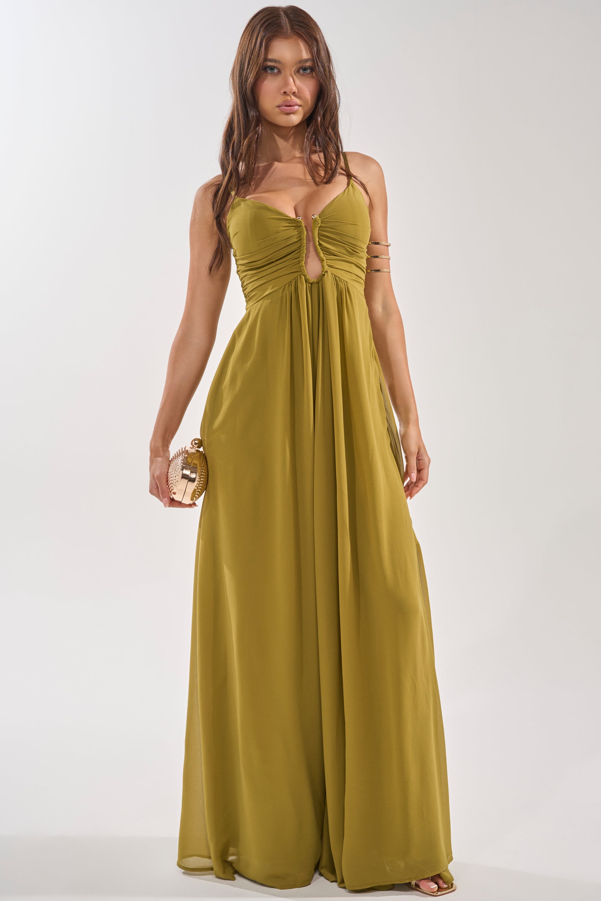 A woman in the LATE NIGHT IN KAUAI JUMPSUIT, an olive wide-leg style with a gathered bodice and keyhole cutout, holds a small gold clutch. She has long wavy brown hair and stands against a plain light background. 0