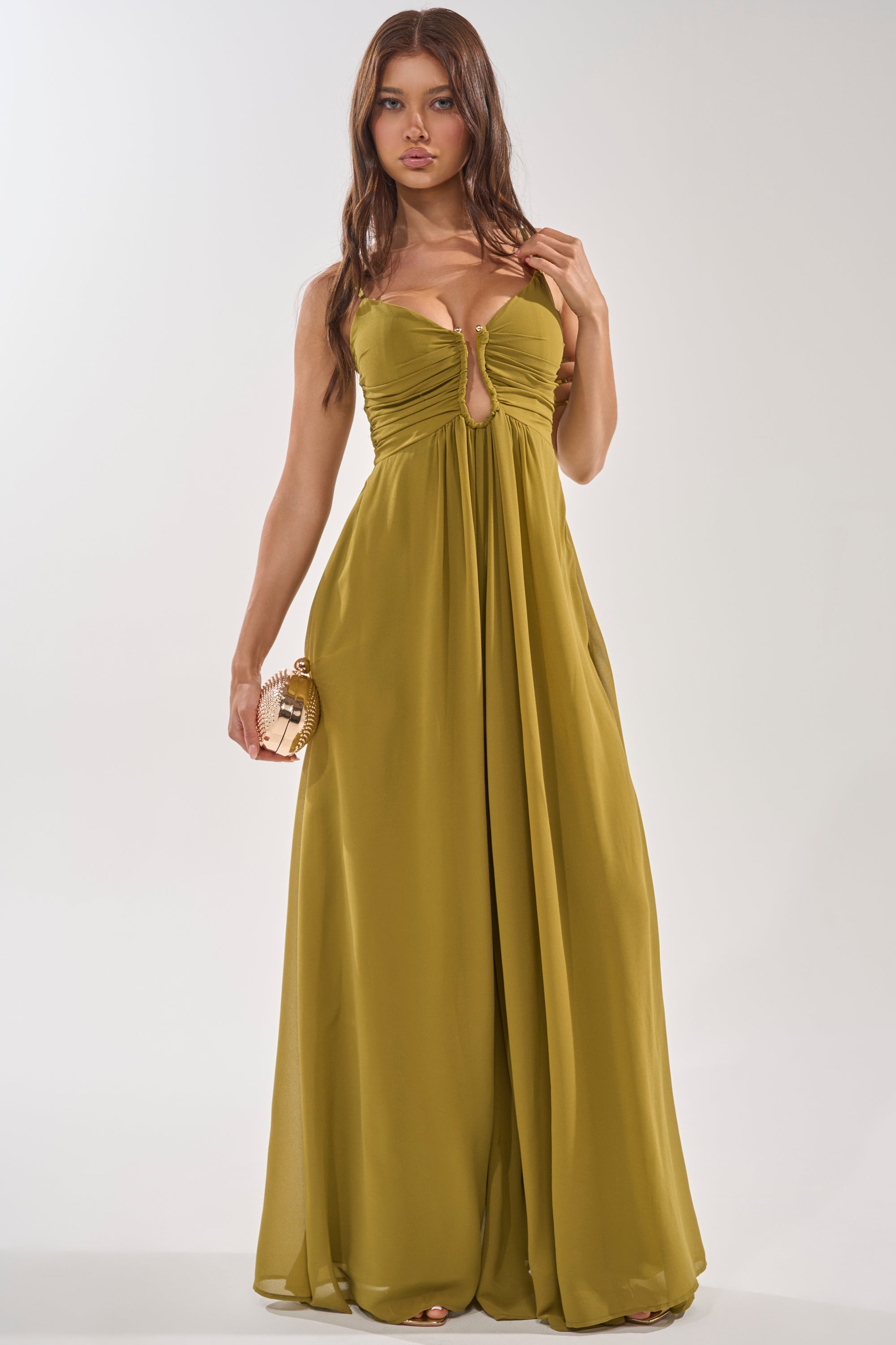 A woman wears the LATE NIGHT IN KAUAI JUMPSUIT, an olive green, floor-length style with a sweetheart neckline, keyhole cutout, and ruched bodice, holding a round clutch as the jumpsuit flows elegantly to the ground. 1