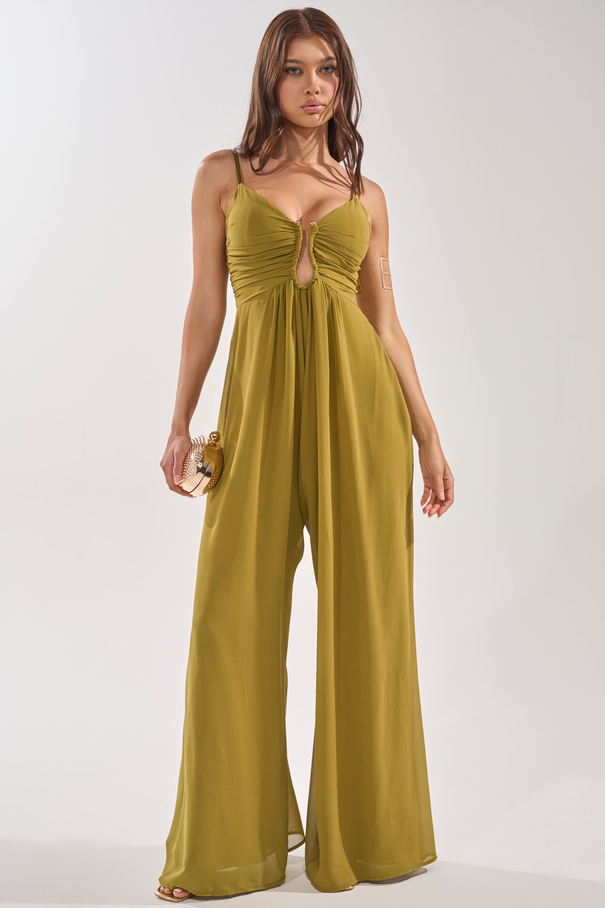 A woman stands against a plain background in the LATE NIGHT IN KAUAI JUMPSUIT, an olive wide-leg style with a ruched keyhole bodice. Perfect for wedding guests, she holds a small round clutch and wears her long hair loose. 2