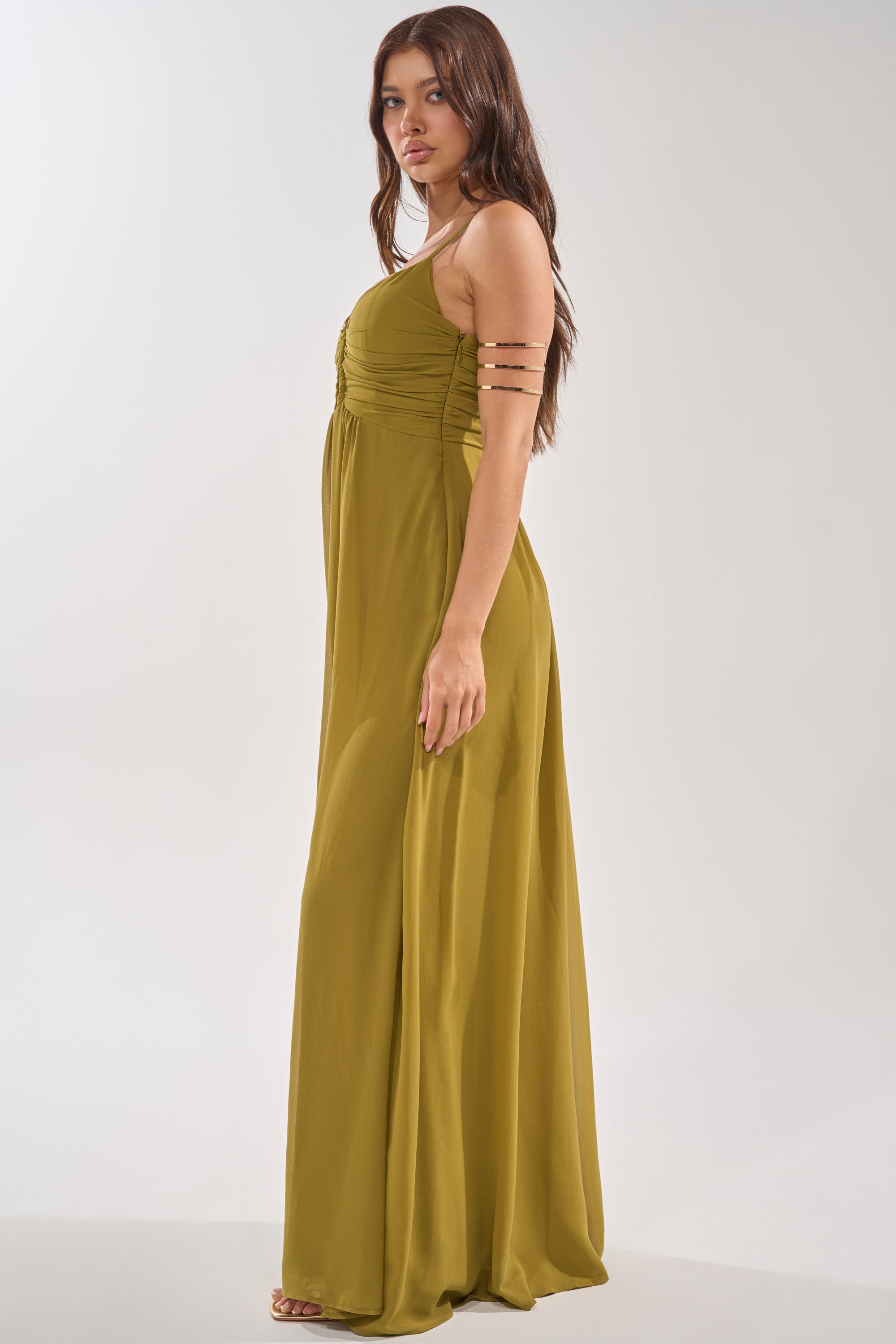 A woman in profile wears the LATE NIGHT IN KAUAI JUMPSUIT, featuring an olive ruched bust, fitted bodice, and wide-leg silhouette. Her long brown hair and gold arm cuff stand out against the simple light background for an elegant look. 3