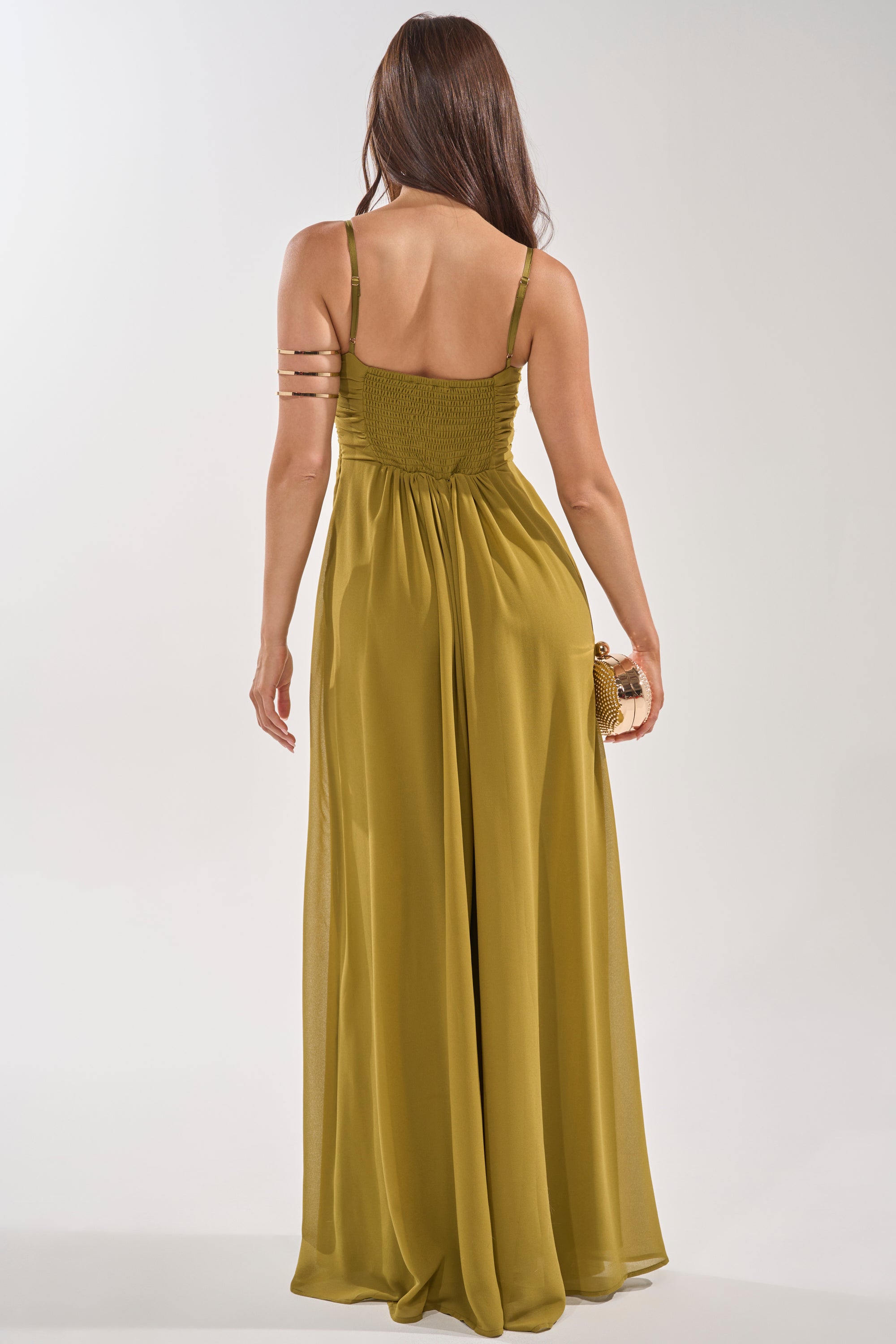 A woman with long brown hair, seen from behind, wears the LATE NIGHT IN KAUAI JUMPSUIT in olive and holds a small clutch. She has a gold arm cuff on her upper left arm. The background is plain and light-colored. 4